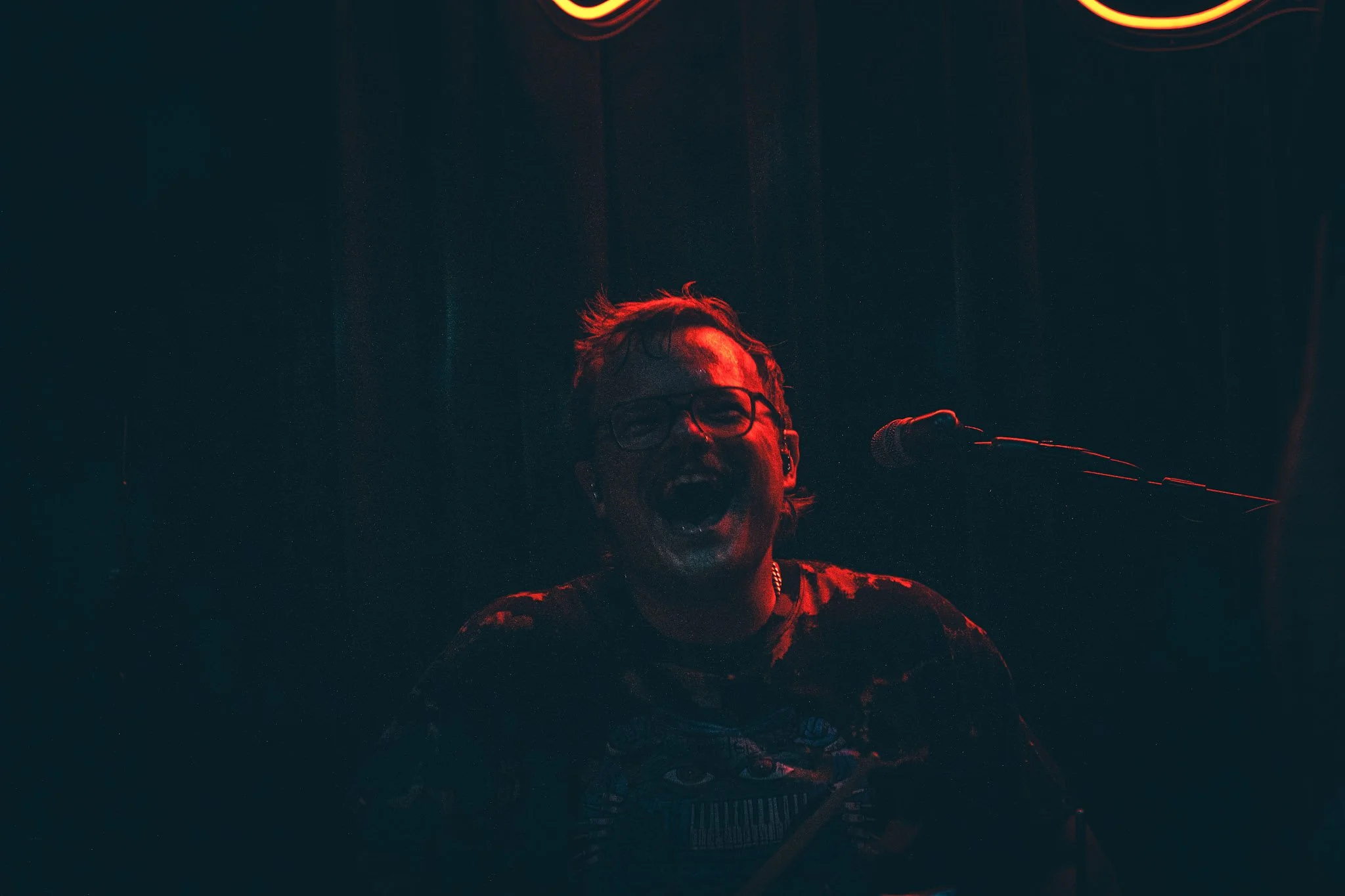 A person with glasses singing into a microphone on a dark stage illuminated by red lighting.