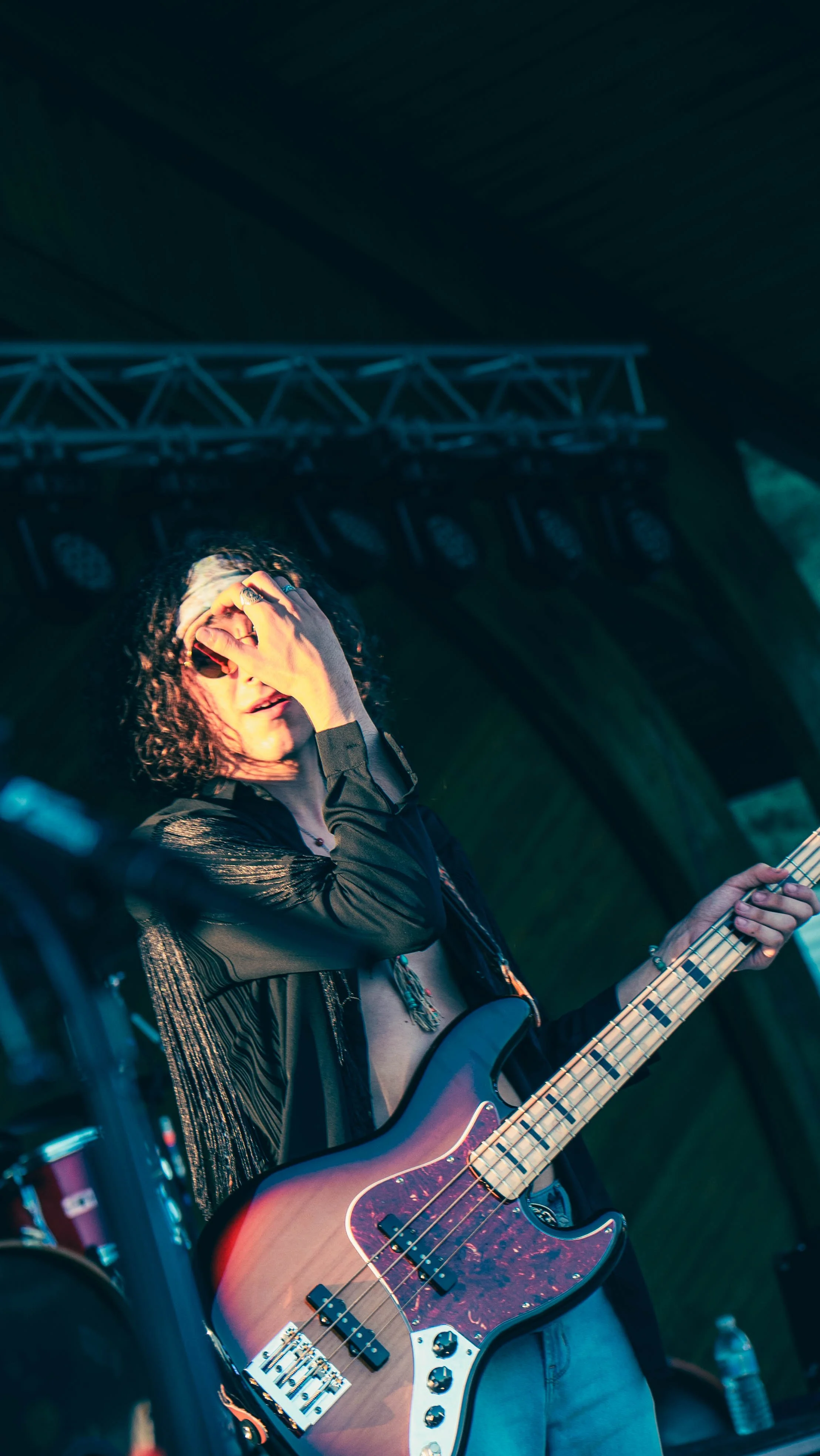 A musician with curly hair and sunglasses playing an electric bass guitar on stage, holding his hand over his face, wearing a black leather jacket, with stage equipment and lighting in the background.