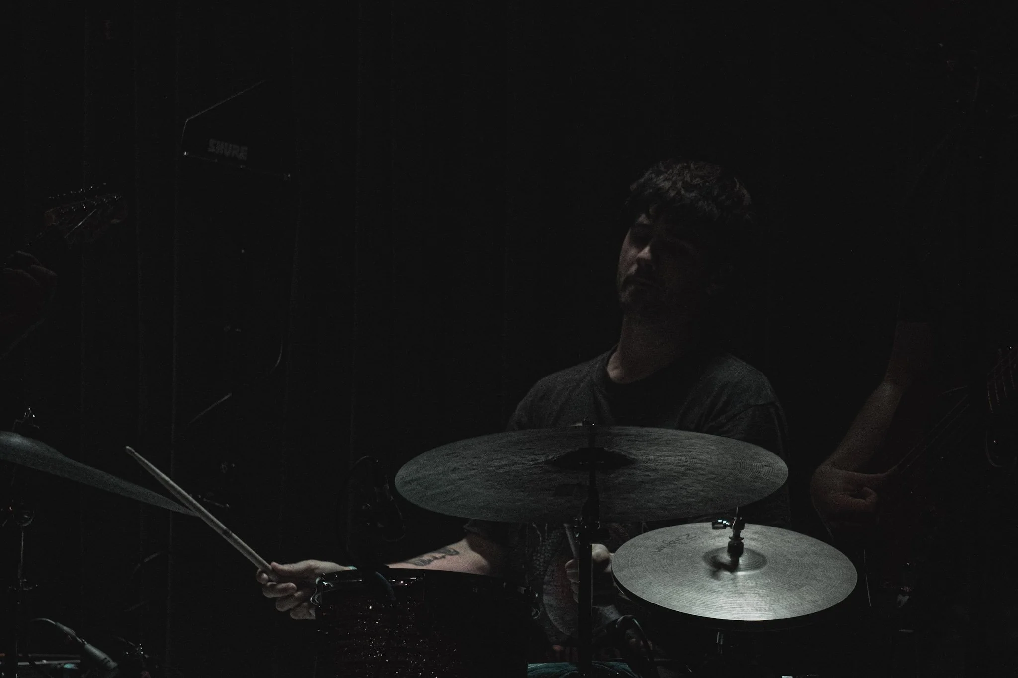 A person playing drums in a dark room with minimal lighting, holding drumsticks, with a cymbal and other drum equipment visible.