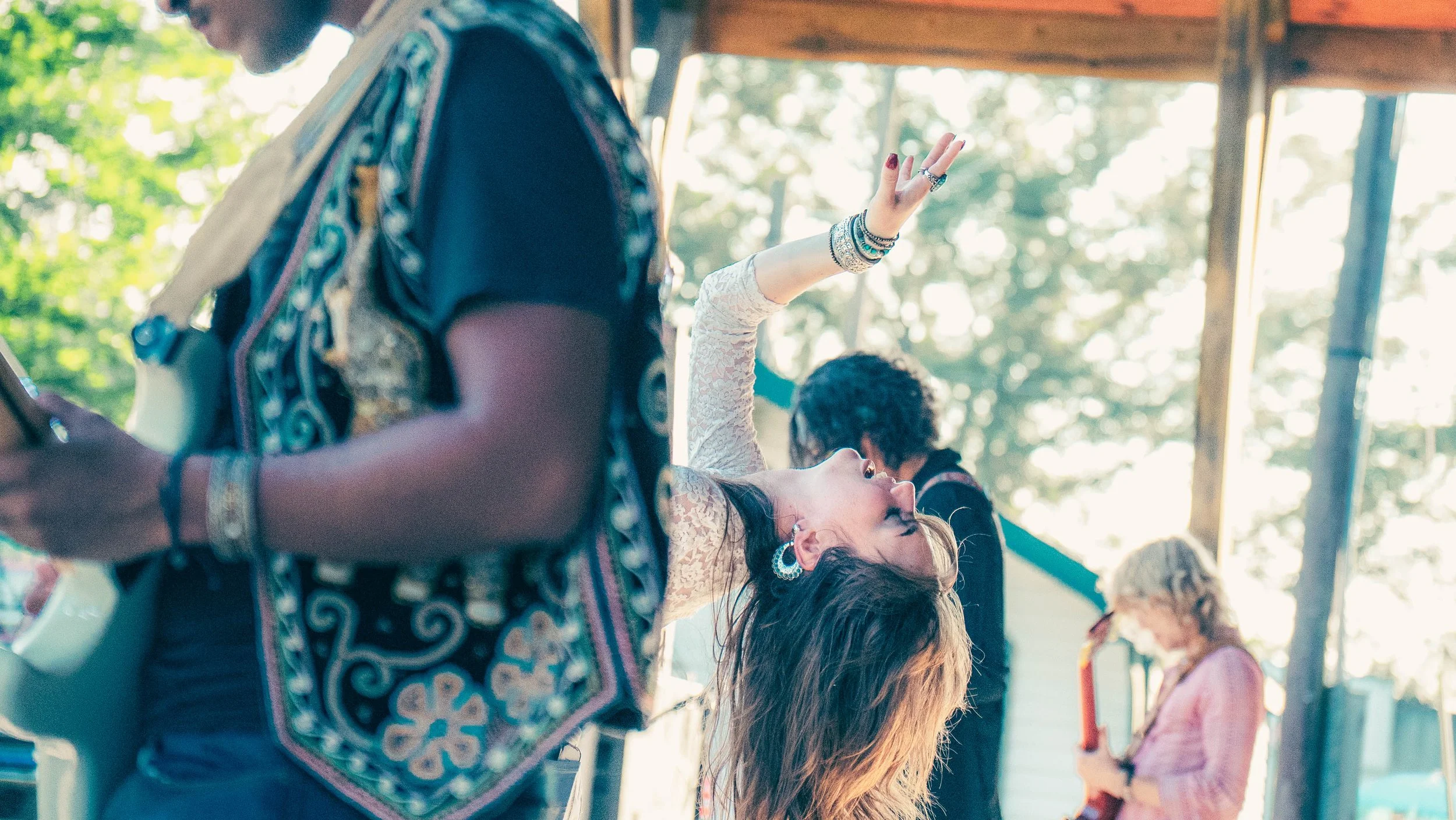 A group of people at an outdoor event, with a focus on a woman in a white lace top reaching up with her hand. The image shows diverse attendees, some wearing glasses and jewelry, with a background of trees and sunlight.