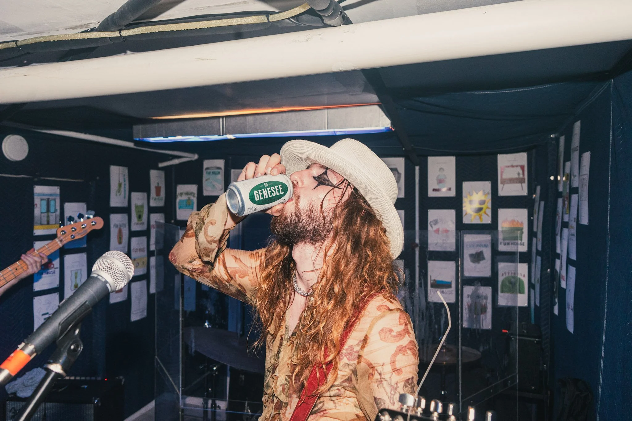 A person with long red hair, a beard, wearing a hat and a camouflage shirt, is drinking from a can of Genesee beer. They are in a room with musical instruments and artwork on the black walls, and a microphone is visible in the foreground.