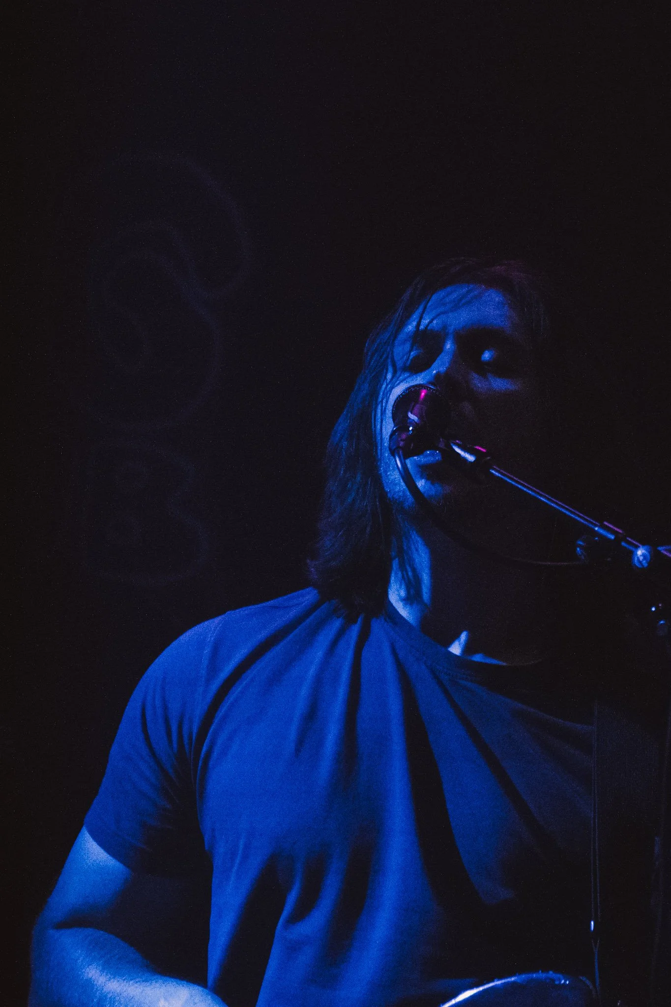 A male musician with long hair singing into a microphone, illuminated by blue stage lighting, wearing a short-sleeved sports shirt.