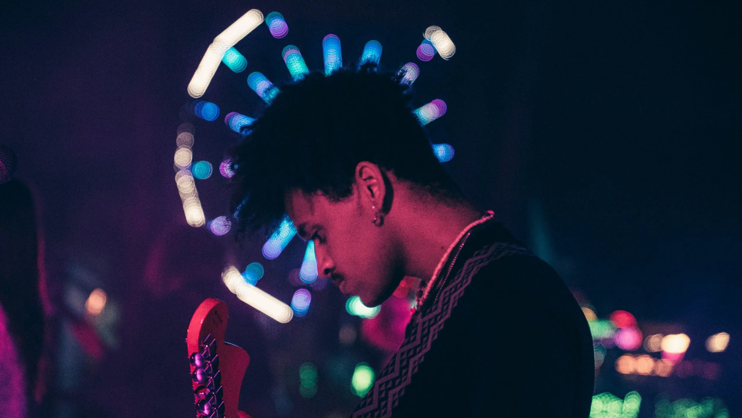 A person with short curly hair looking down surrounded by colorful blurred lights, possibly at a nightclub or festive setting.