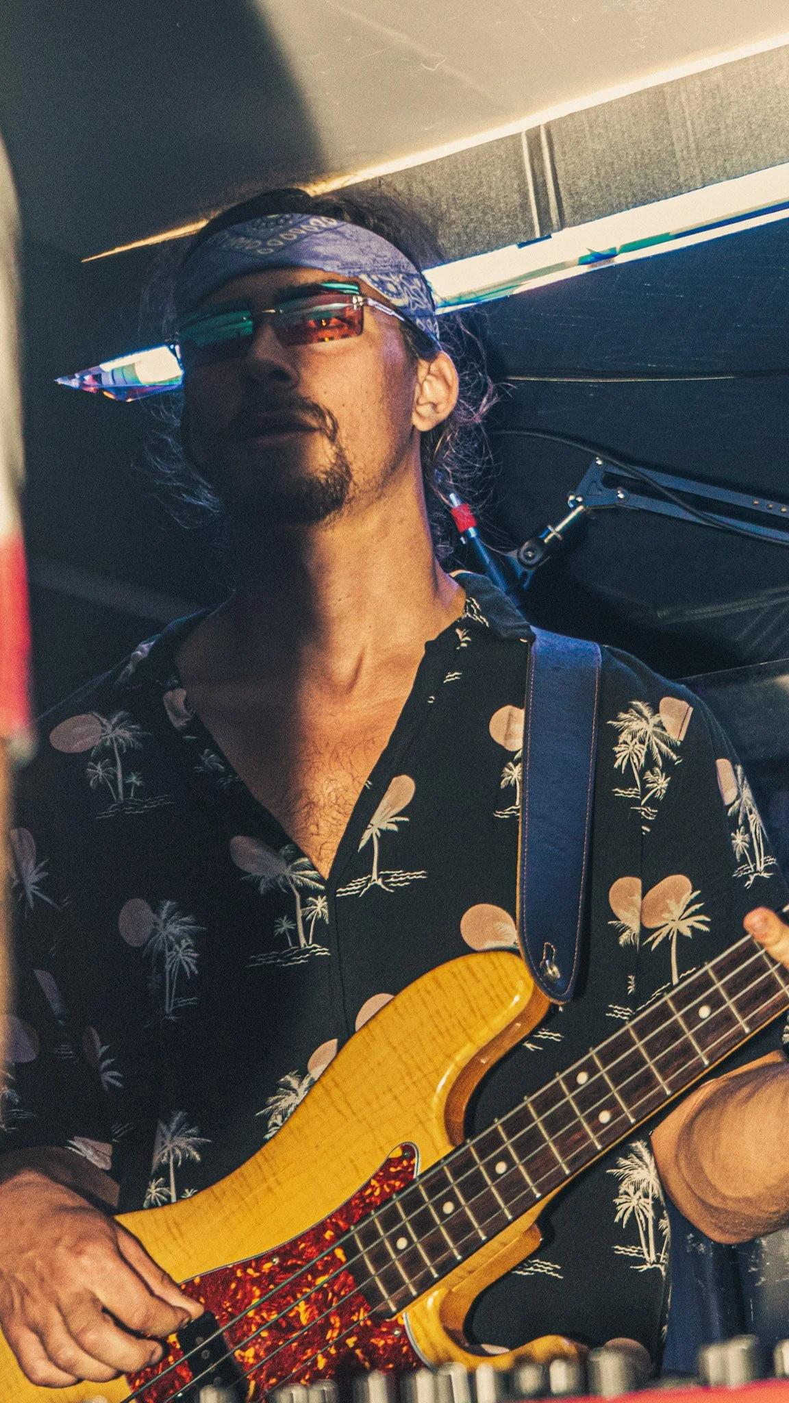 A man wearing sunglasses, a blue bandana, and a black Hawaiian shirt with palm trees and moon pattern, playing an acoustic guitar with a red and black tortoiseshell pickguard.
