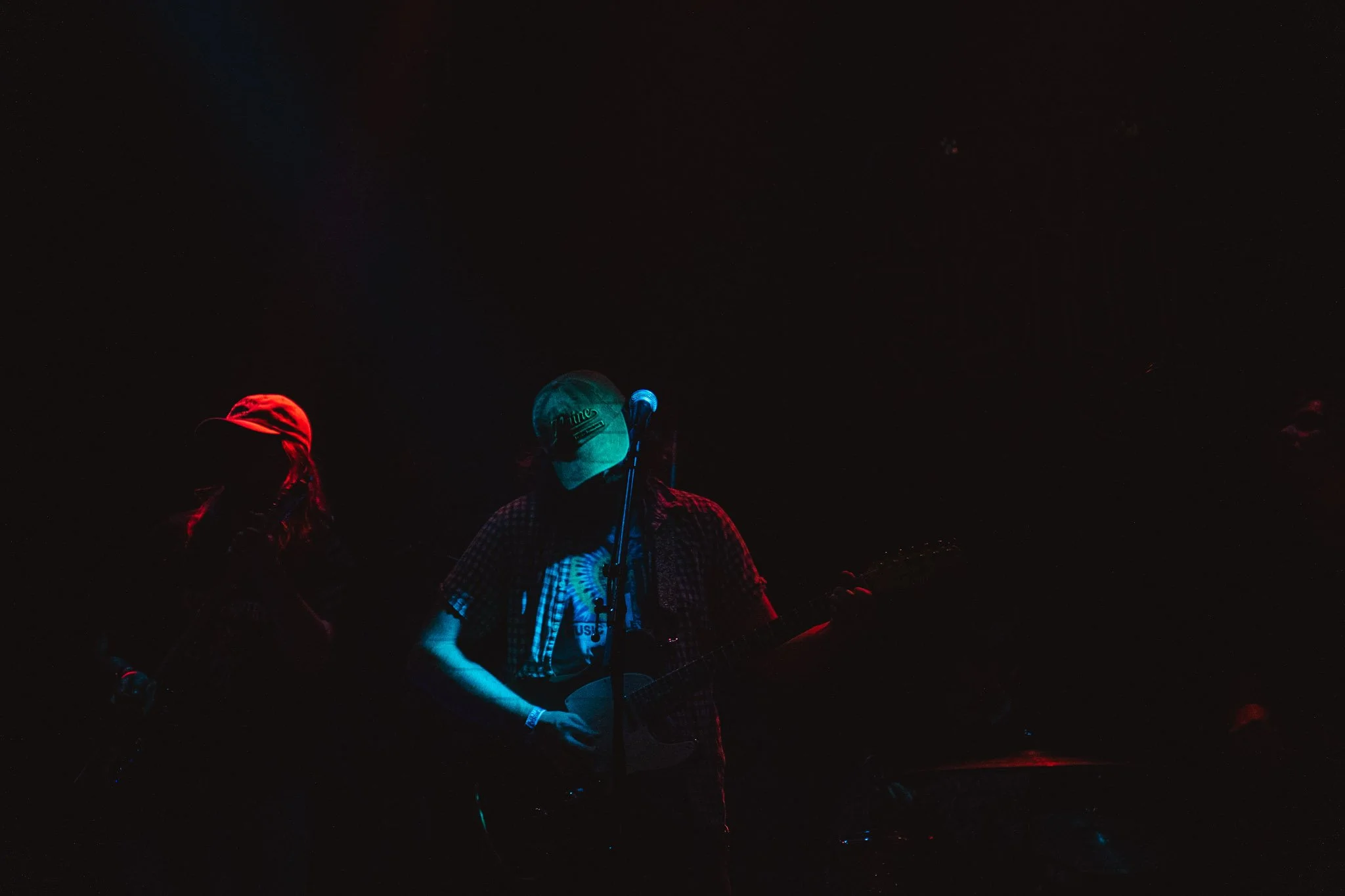 A dark scene with a person wearing a baseball cap and plaid shirt playing a guitar, illuminated by faint red and blue lights, with other people partially visible in the background.