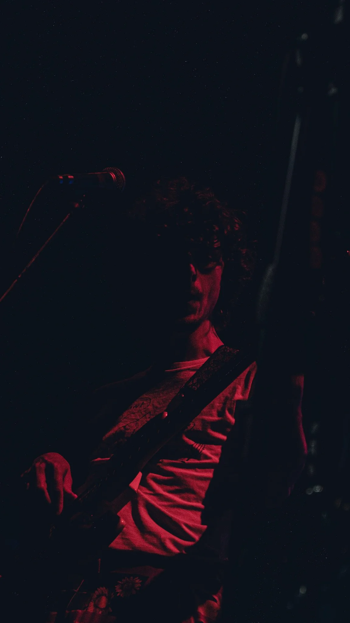 A person with curly hair playing a guitar on a dark stage illuminated by red lighting, with a microphone stand nearby.