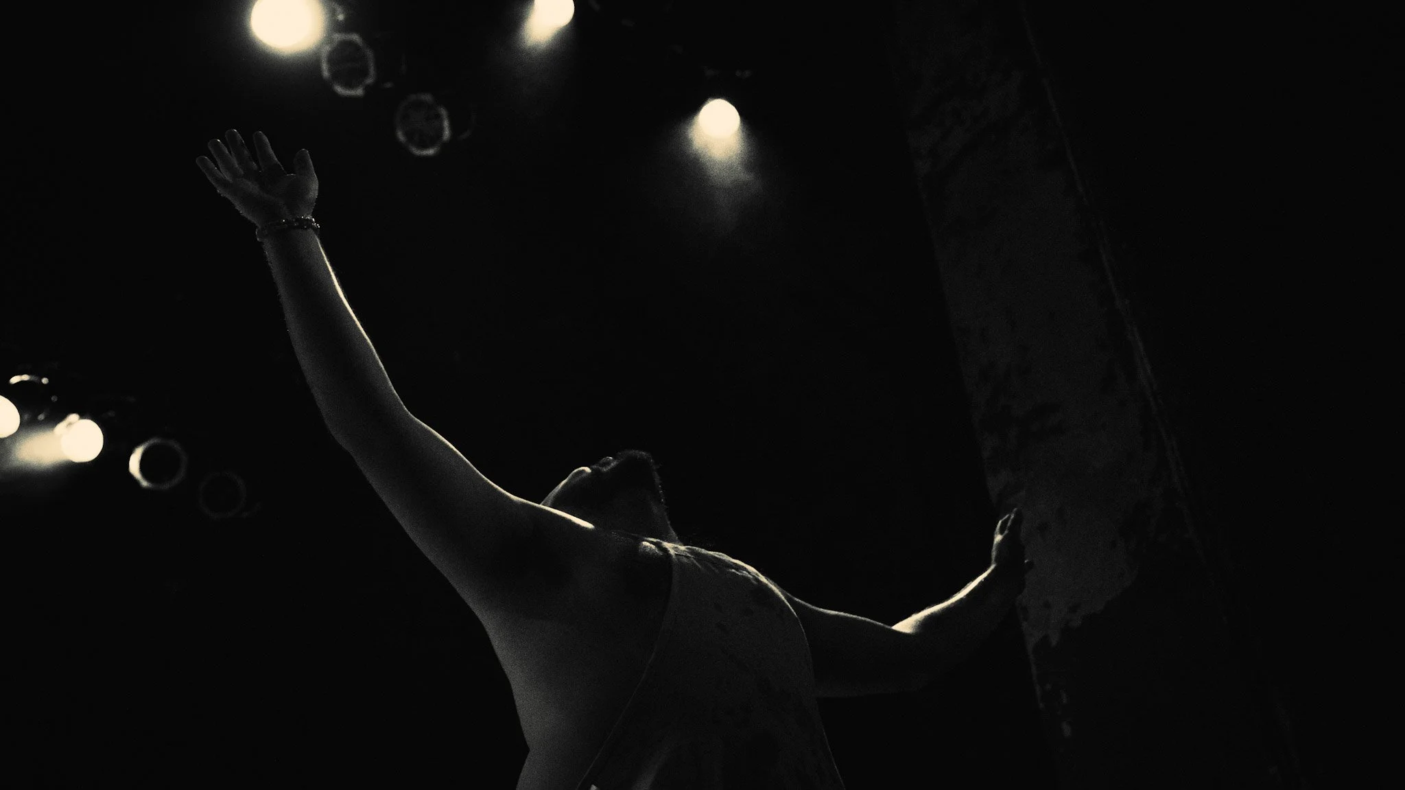 A performer on stage with their arm raised, illuminated by bright stage lights against a dark background.