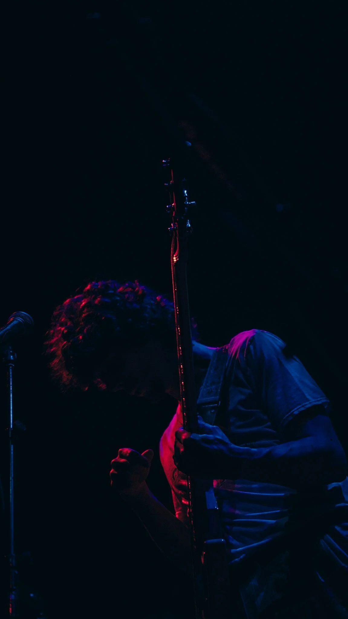 A musician on stage playing a guitar with stage lighting highlighting him in a dark background.