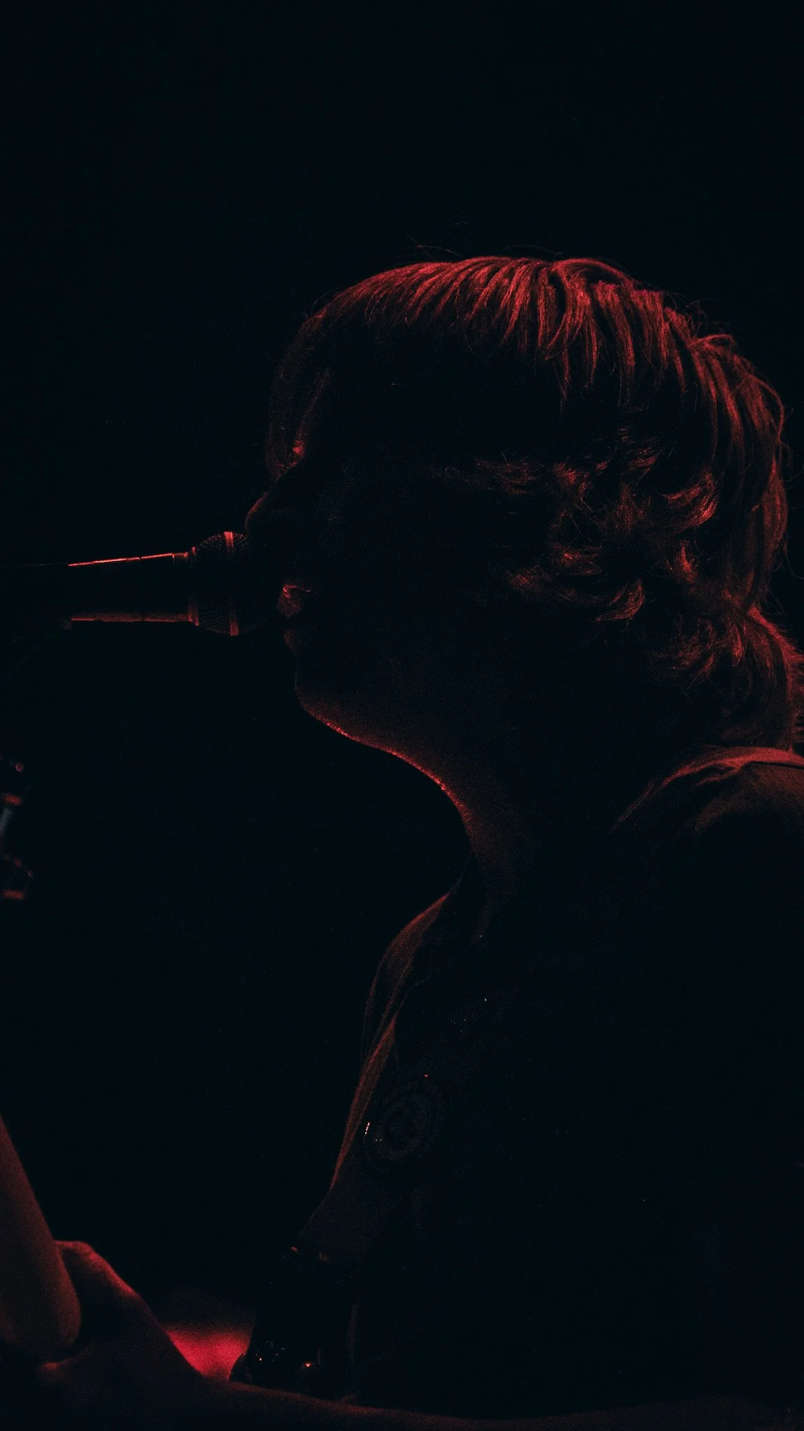 Side profile of a person with curly hair singing into a microphone, illuminated by red lighting in a dark background.