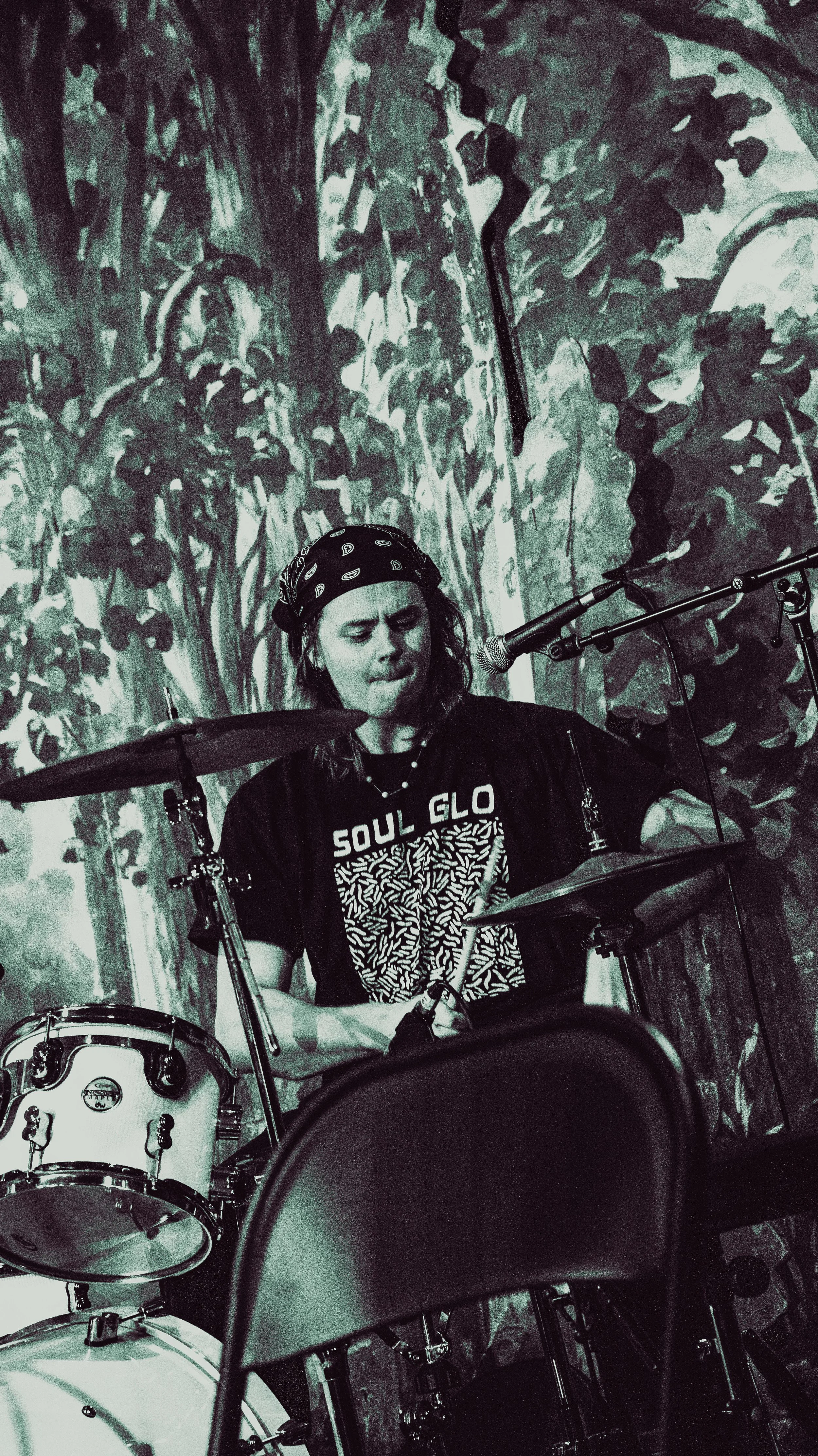 A person with long hair wearing a bandana, playing a drum set in front of an abstract mural background.