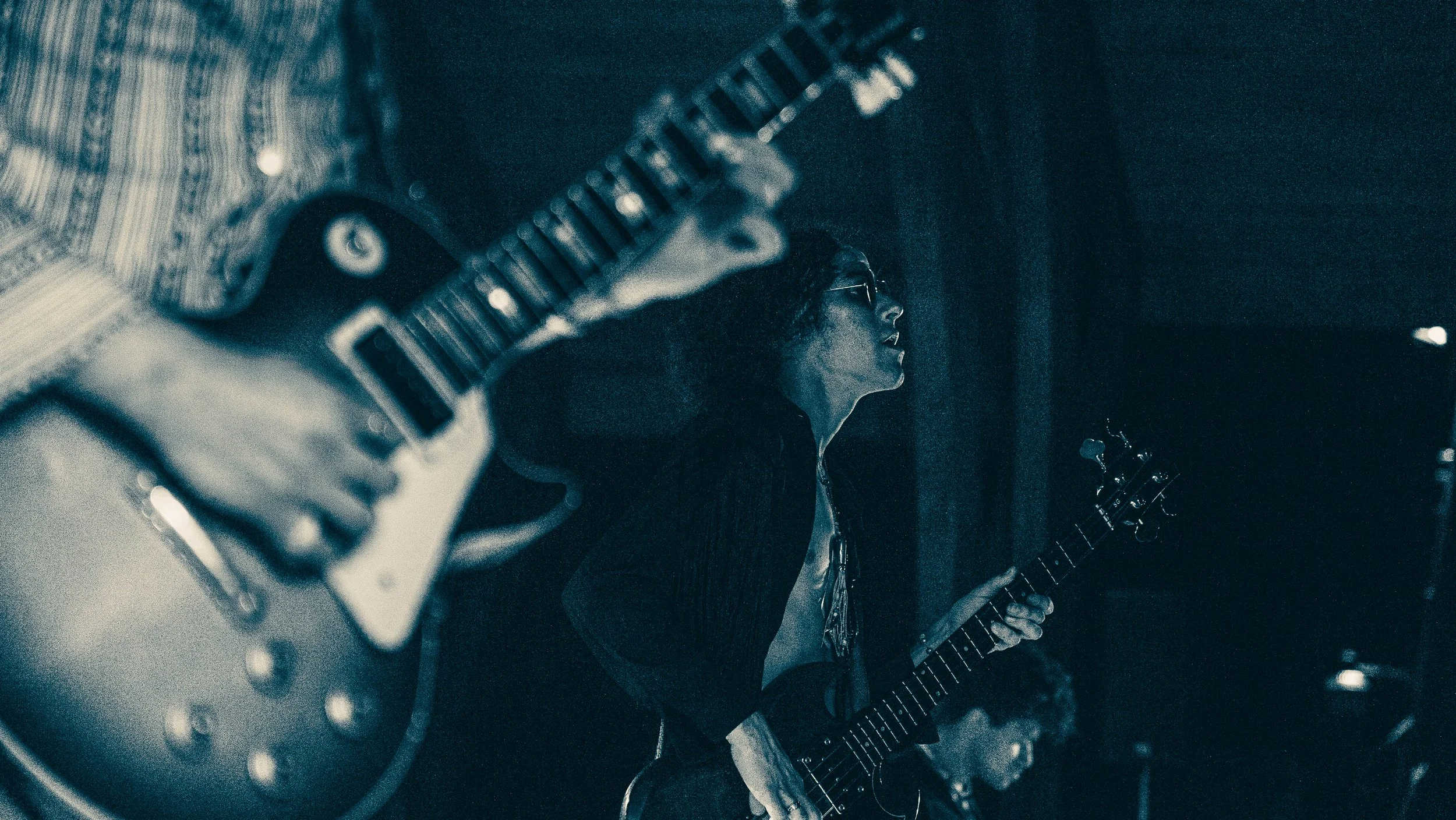 A woman with glasses playing an electric guitar in a dark room, with a vintage radio or appliance visible in the foreground.