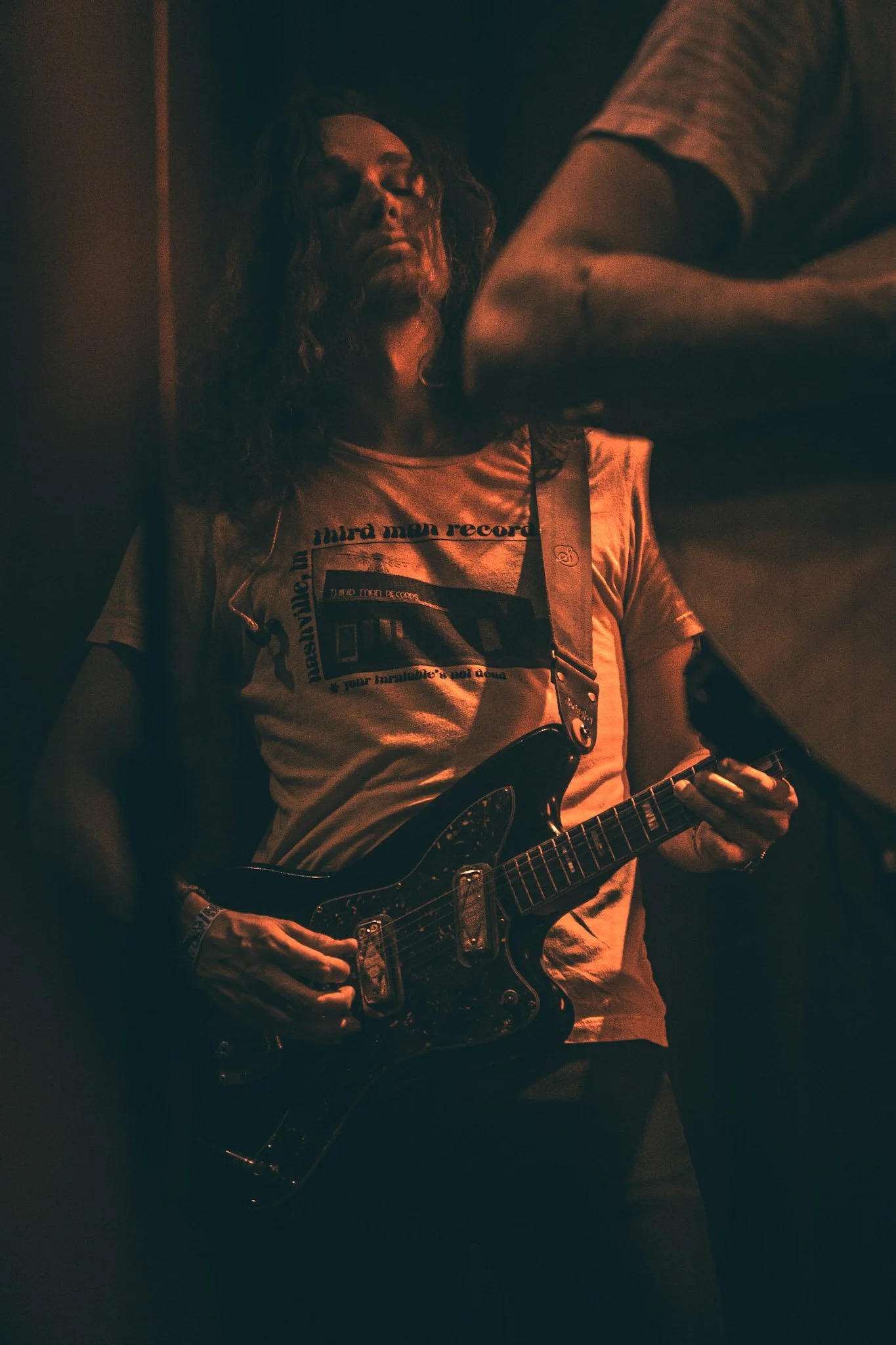A person playing an electric guitar in a dimly lit environment, wearing a light-colored T-shirt with text and graphics.