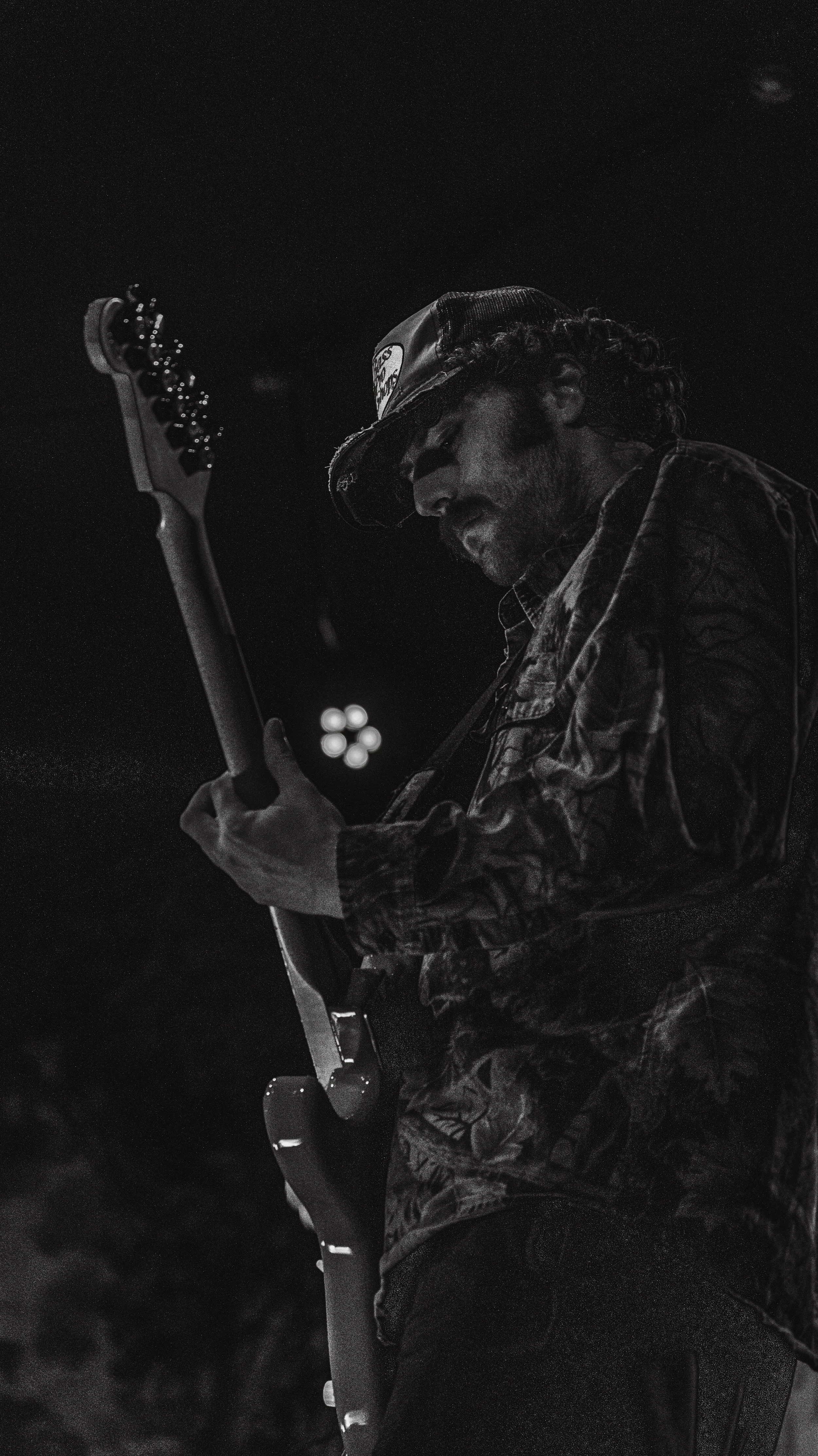 A man wearing a camouflage jacket and a cap, playing an electric guitar, with a dark background and bright stage lights.