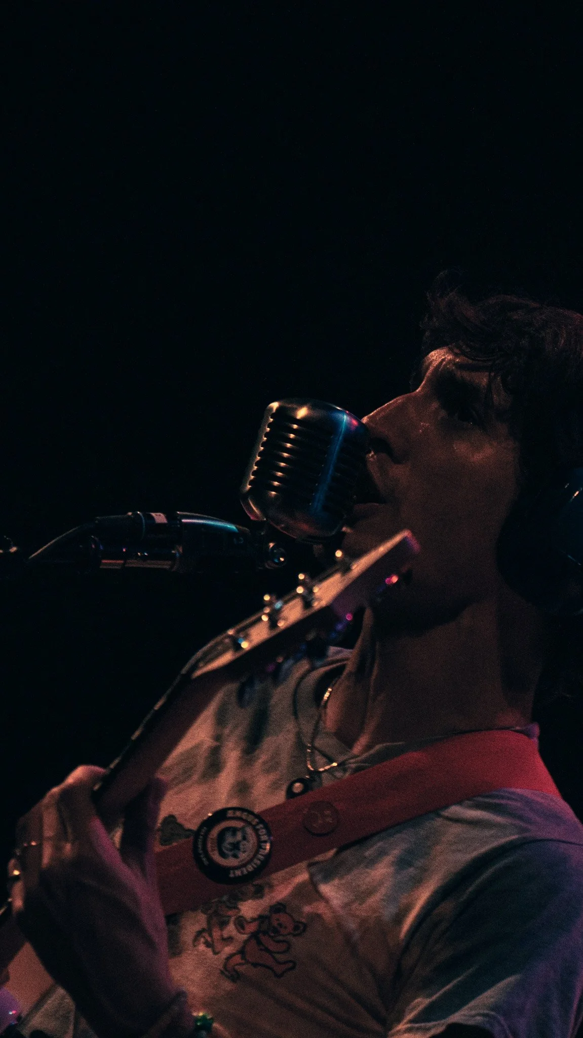 A young man singing into a vintage microphone in a dark setting, wearing a baseball cap and casual clothing.