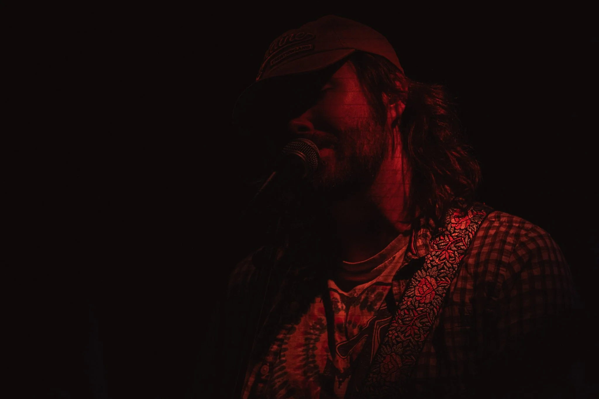Man with long hair and beard singing into a microphone, wearing a baseball cap and a plaid shirt, illuminated by red lighting against a dark background.