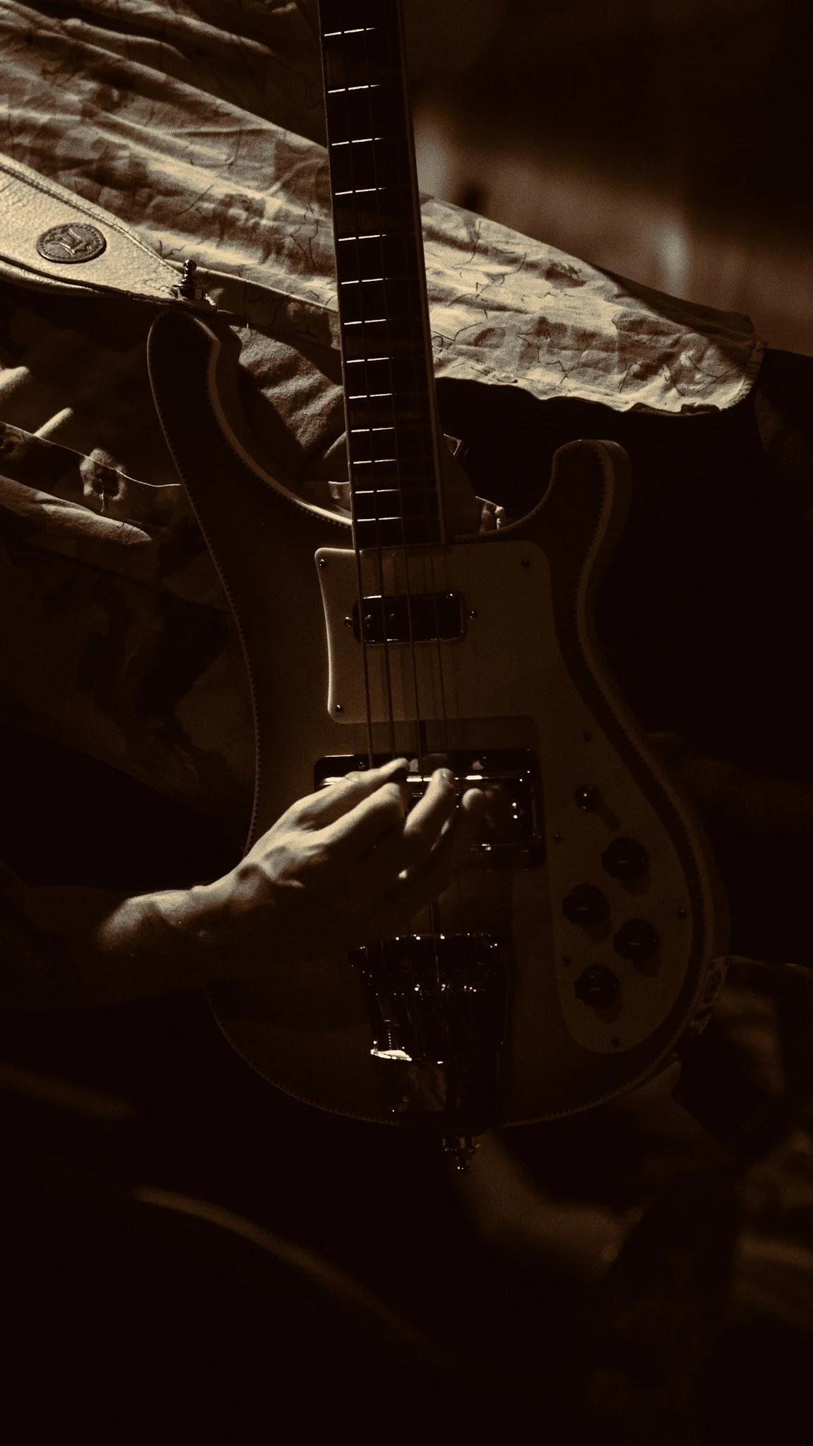 Close-up of a person playing an electric guitar in a dimly lit room. The guitar is cream-colored with black knobs, and the person's hand is on the strings near the bridge. The background includes a patterned cloth on a table and a guitar strap with a