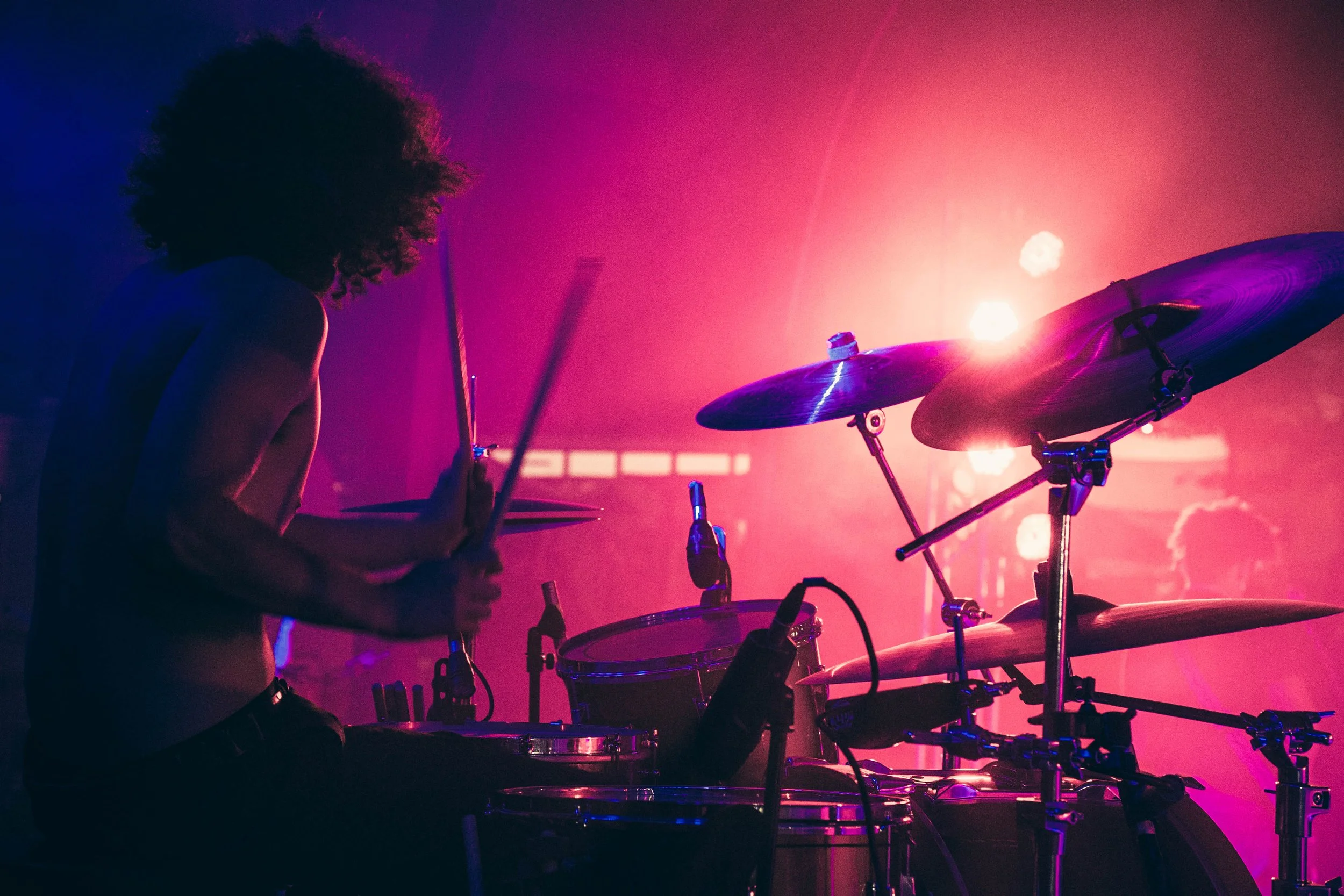 A silhouette of a person with curly hair playing a drum set on a stage illuminated with colorful pink and purple lighting.
