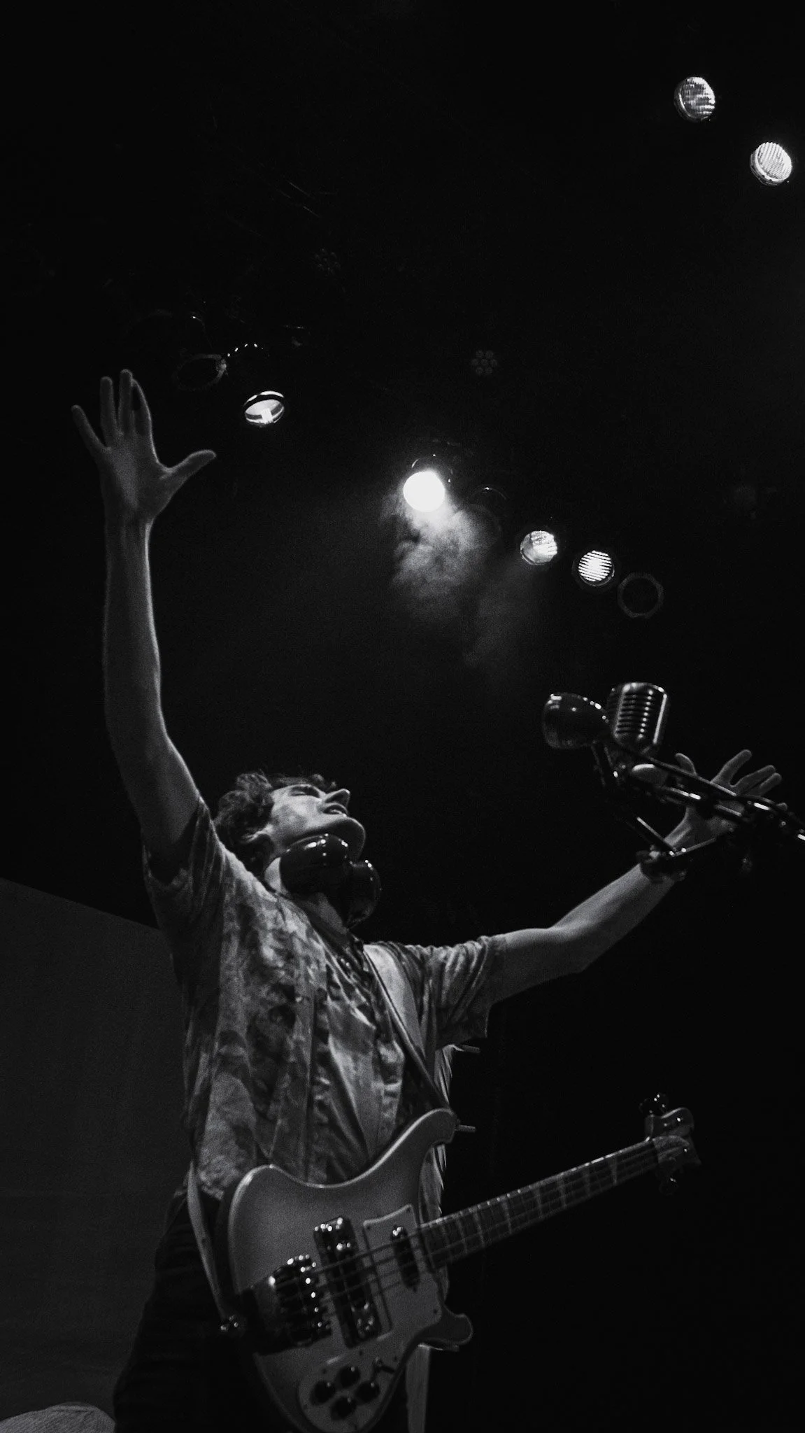 A woman with short hair, wearing glasses and headphones, stands on stage with her hand raised and a guitar around her neck, under stage lighting, with a microphone in front of her.