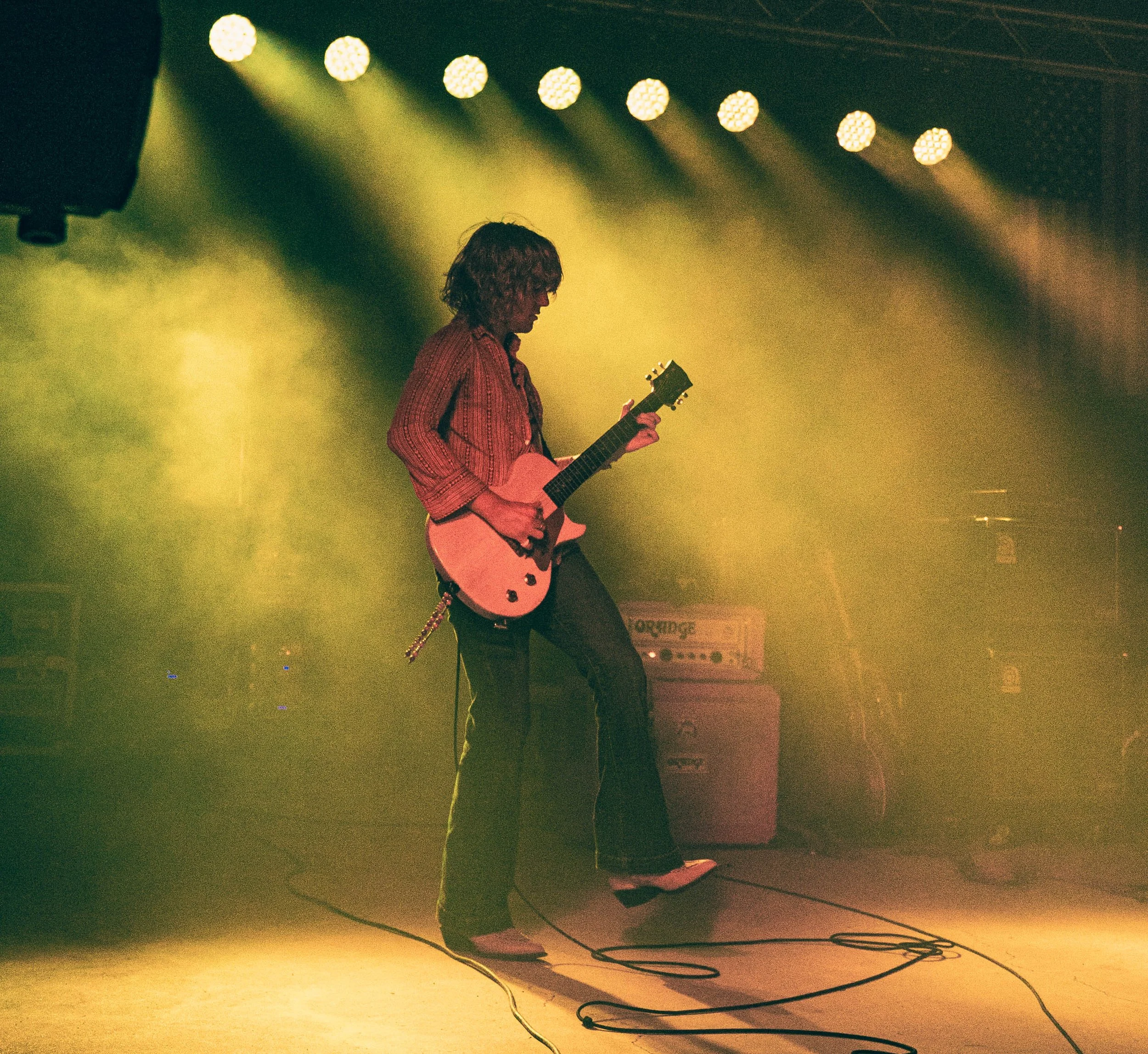 A person playing an acoustic guitar on a stage with greenish-yellow lighting, performing in front of a backdrop with stage equipment and wiring visible on the floor.