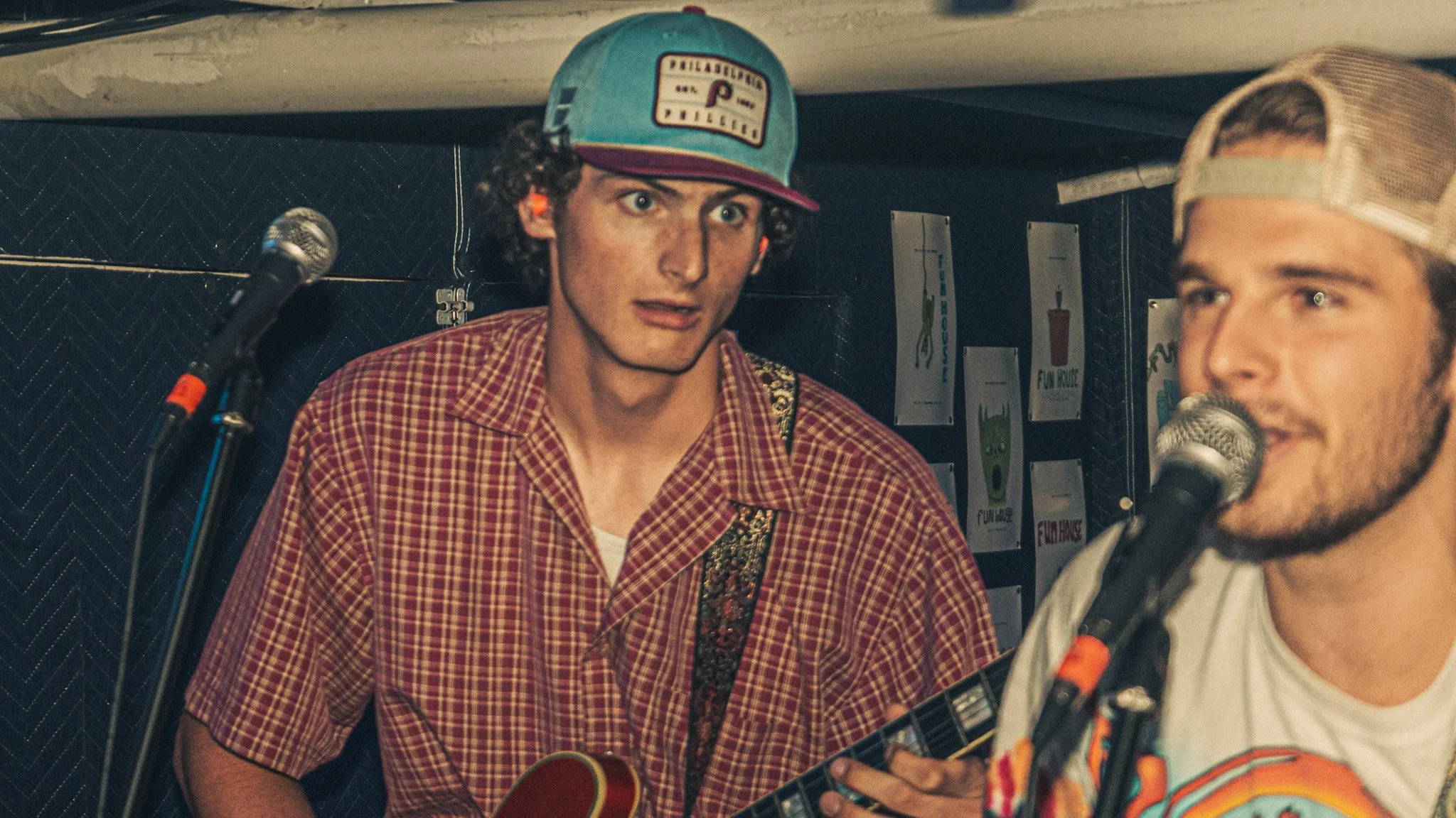 Two men performing with microphones, one playing guitar, in a recording studio or small stage, with posters on the wall.