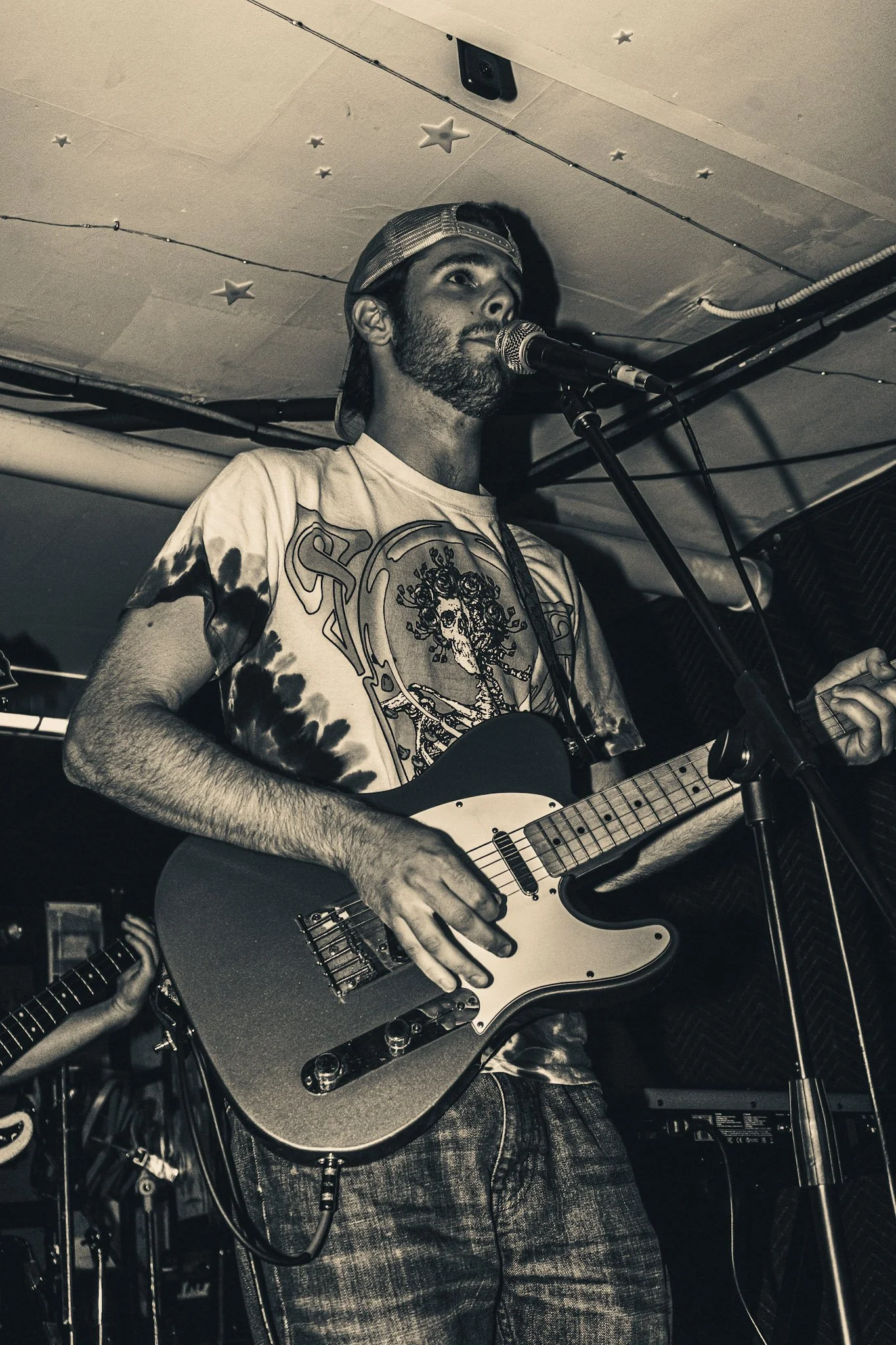 A man playing an electric guitar and singing into a microphone. He is wearing a baseball cap backwards and a graphic t-shirt. The background shows string lights and a speaker.