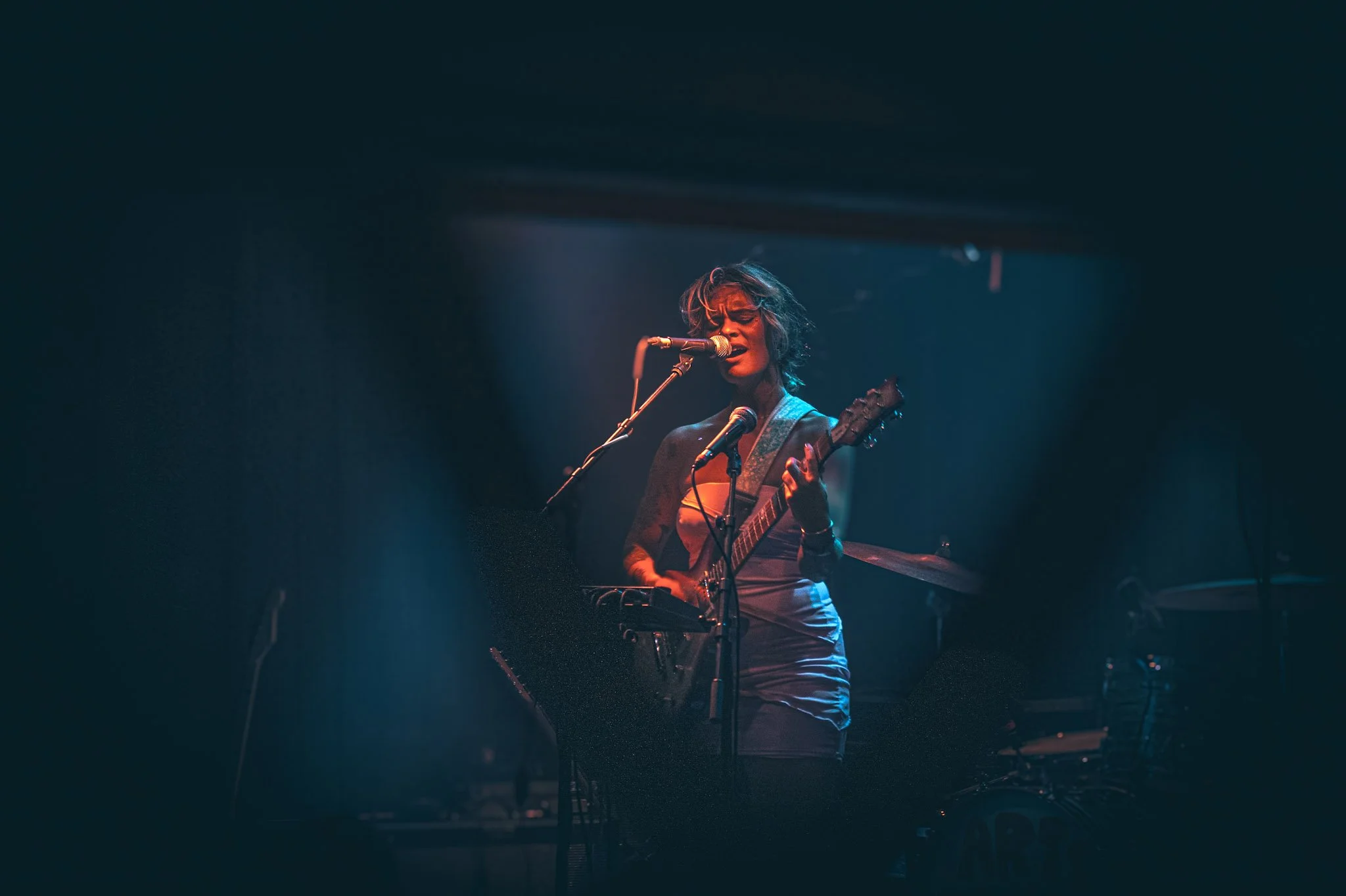 A female musician performs on stage with a guitar and singing into a microphone, lit by stage lights.