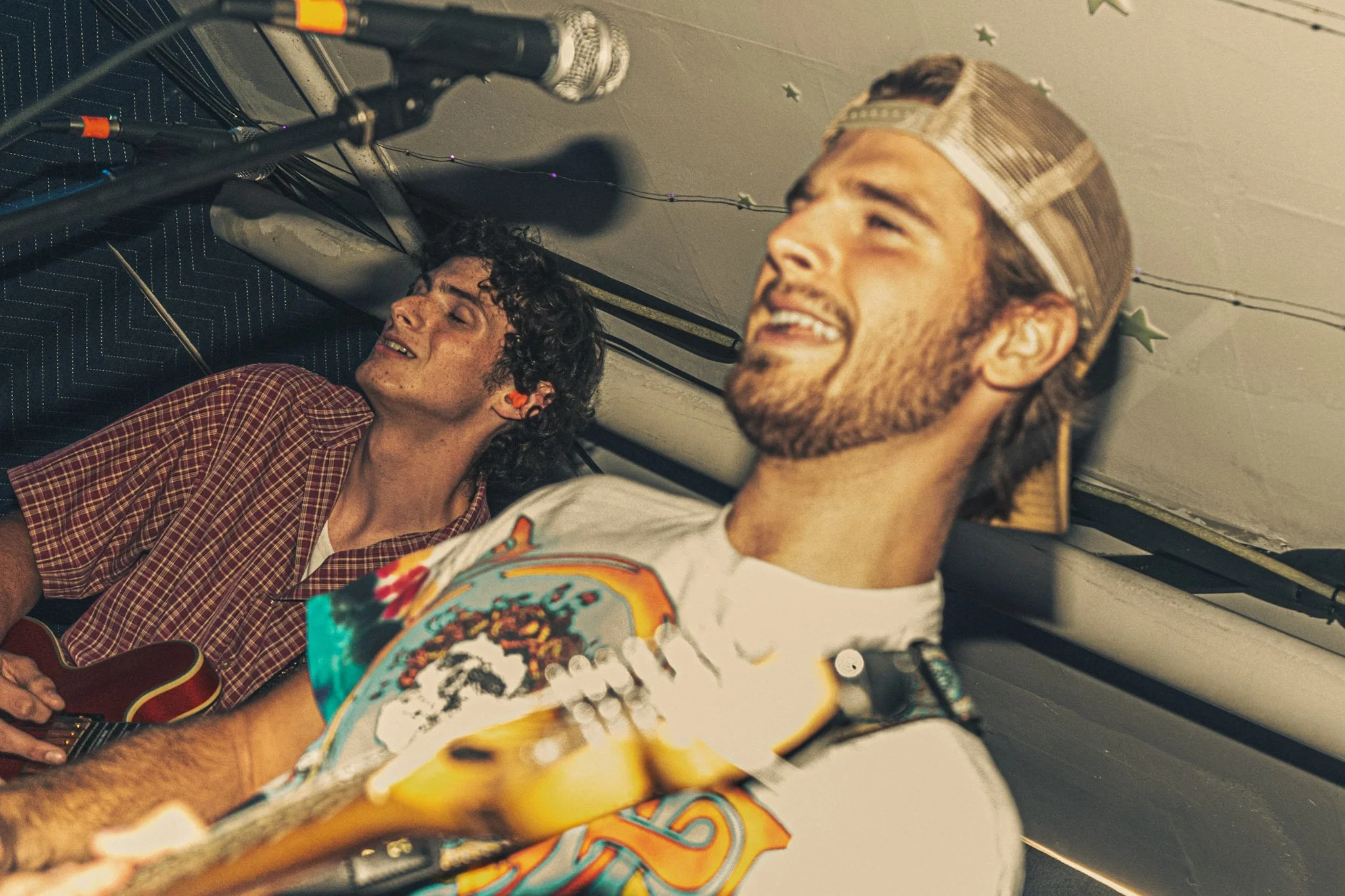 Two men lying on the floor, smiling and relaxed. One has curly hair, wearing a red checkered shirt, holding a guitar. The other has a beard, wearing a colorful graphic T-shirt and a metallic cap.