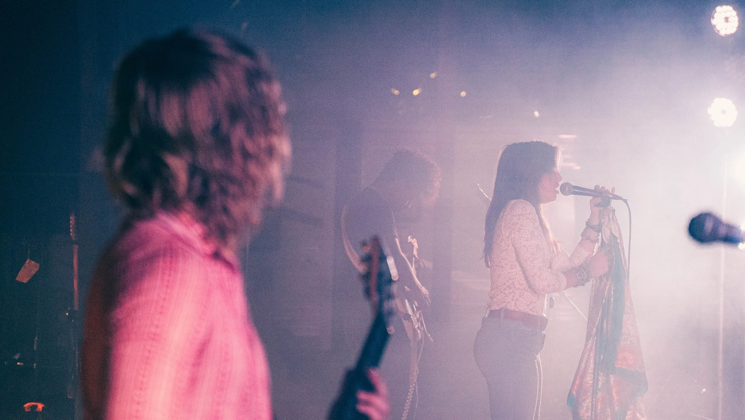A woman sings into a microphone on stage with band members playing instruments in the background, under stage lighting.