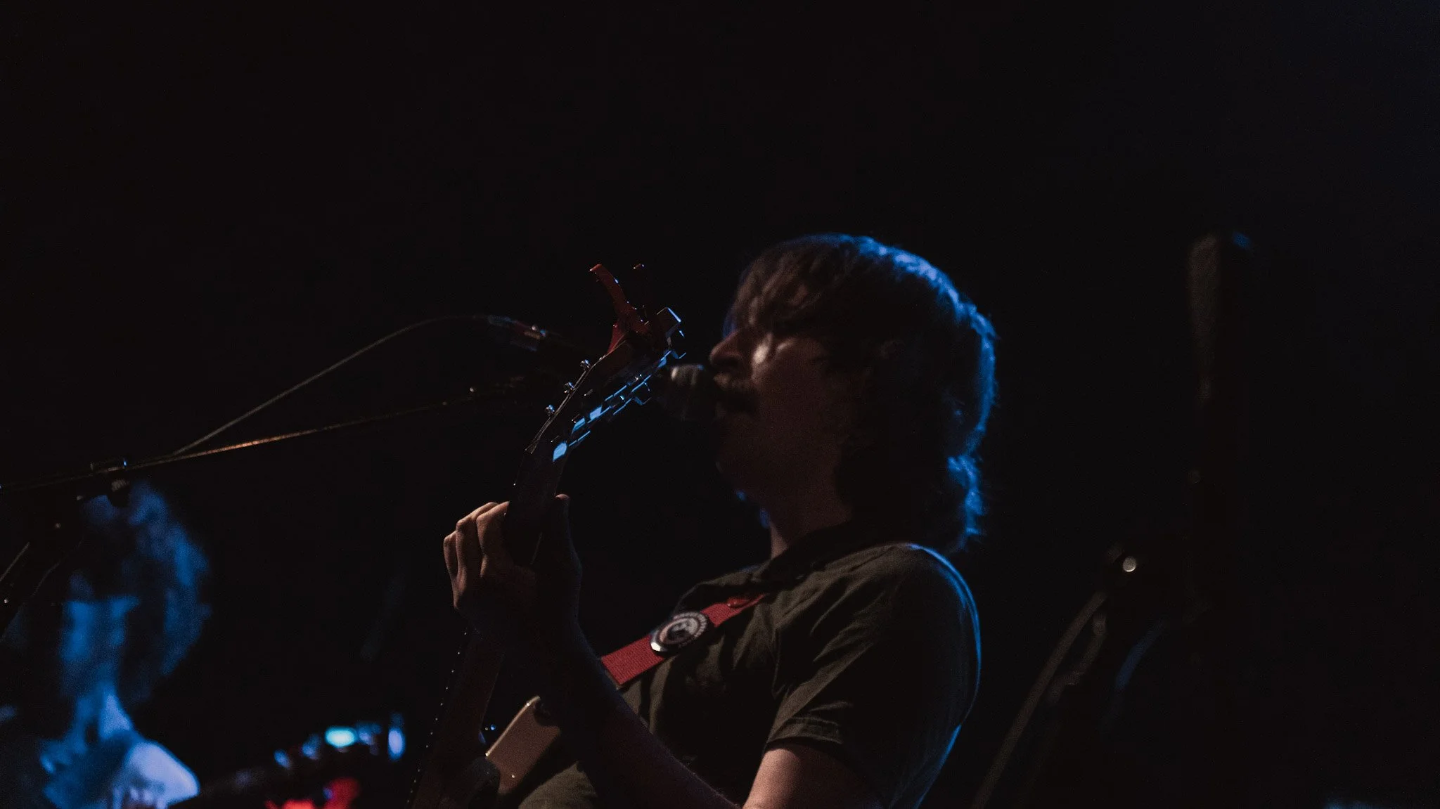 A person with short hair is singing or speaking into a microphone on a dark stage, with blue and red lighting reflections.