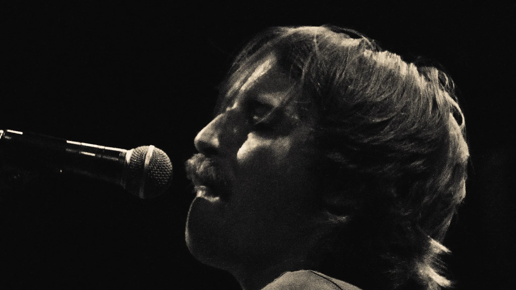 A person singing into a microphone, with light-colored hair, in a dark setting.