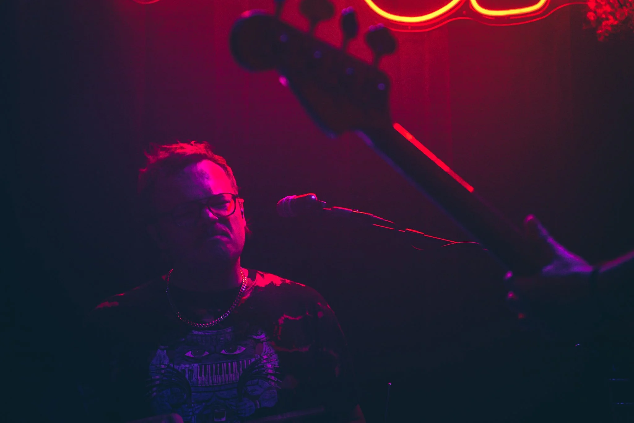 A person with glasses and a patterned shirt playing a keyboard on stage under red and purple neon lights.