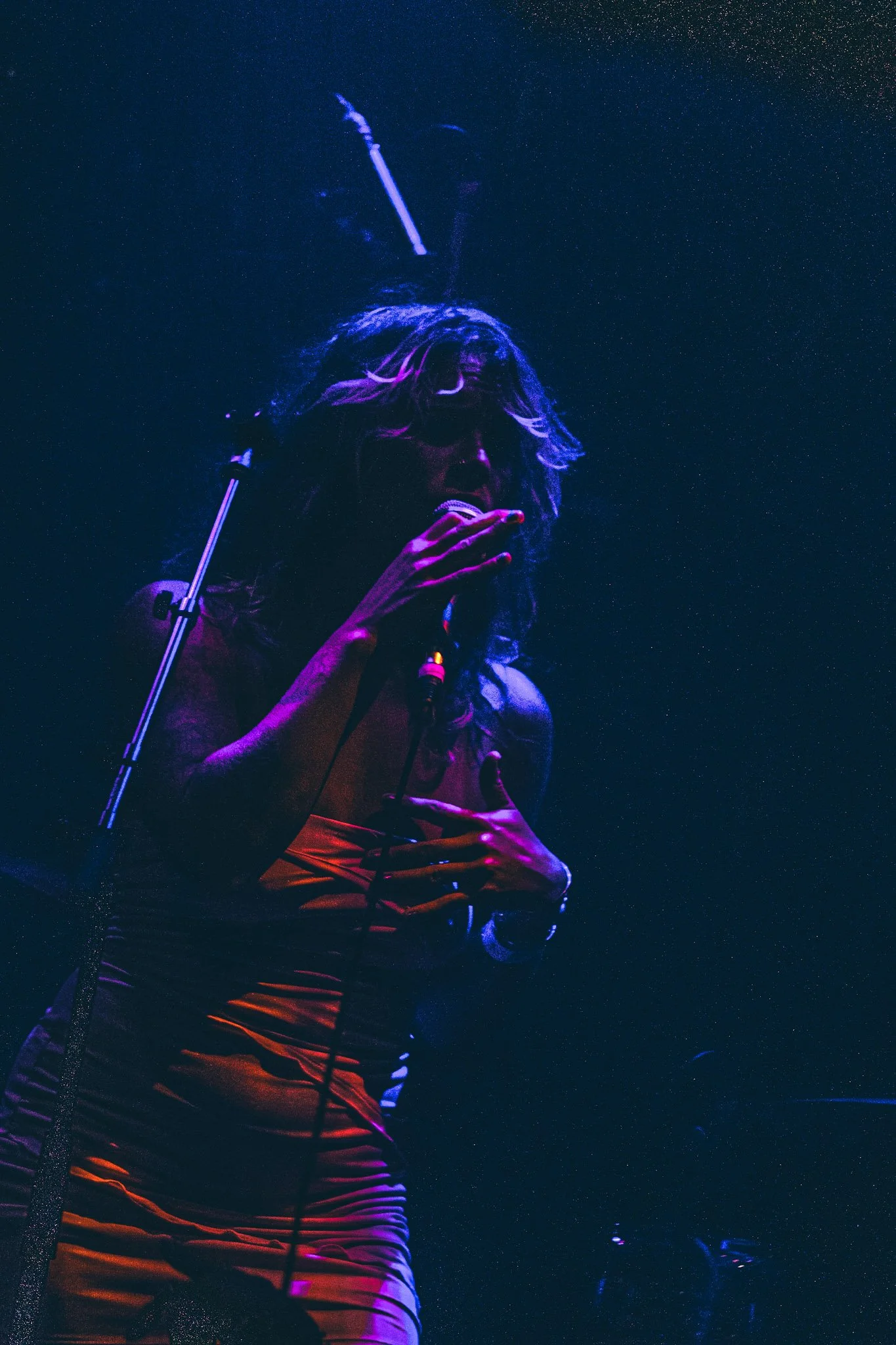 A female singer performs under a starry night sky. She has long, curly hair and is wearing an orange and black striped dress. She is holding a microphone and appears to be singing passionately.