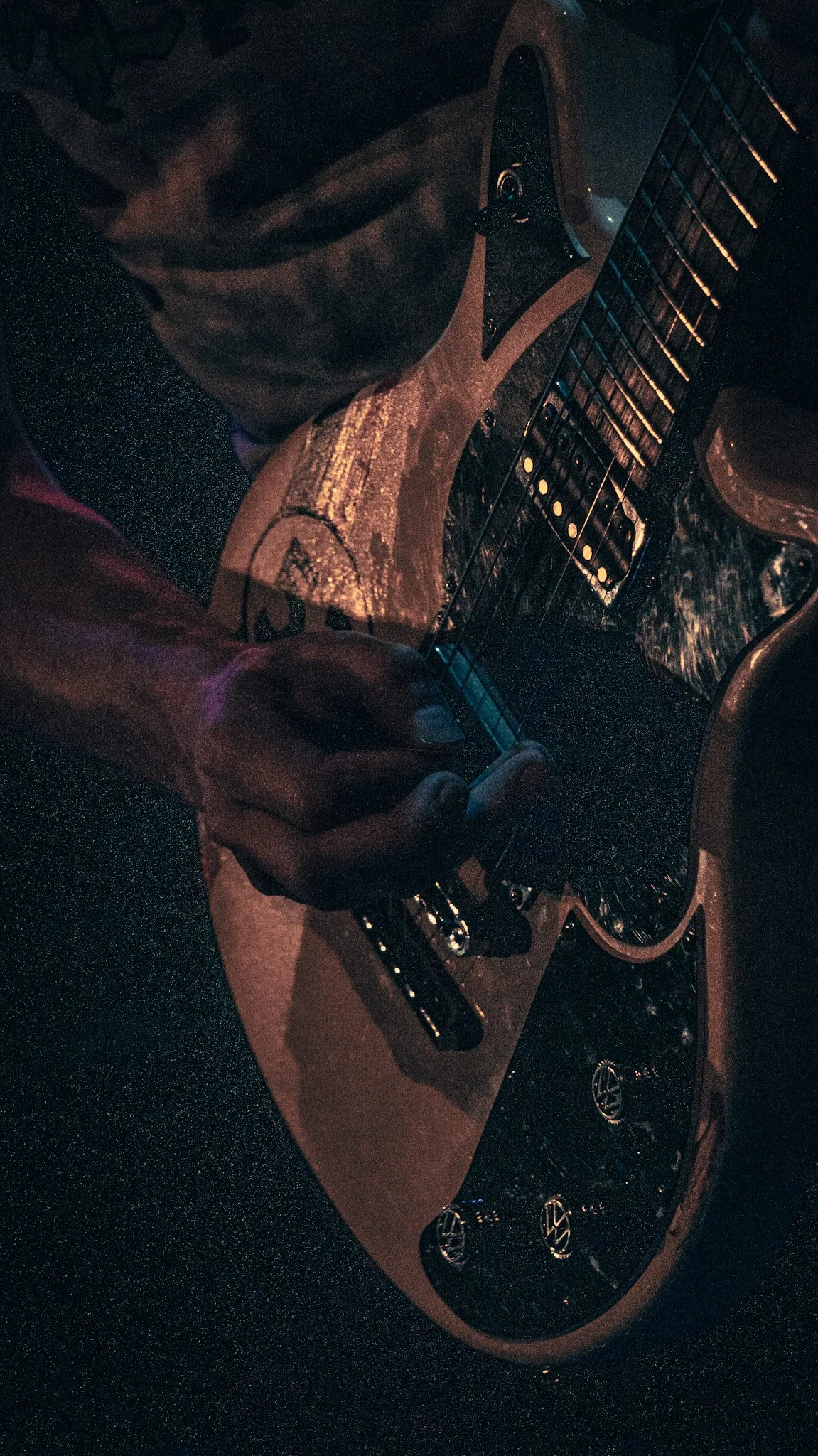 Close-up of a person playing an electric guitar in a dimly lit environment, with the focus on their hand on the fretboard.