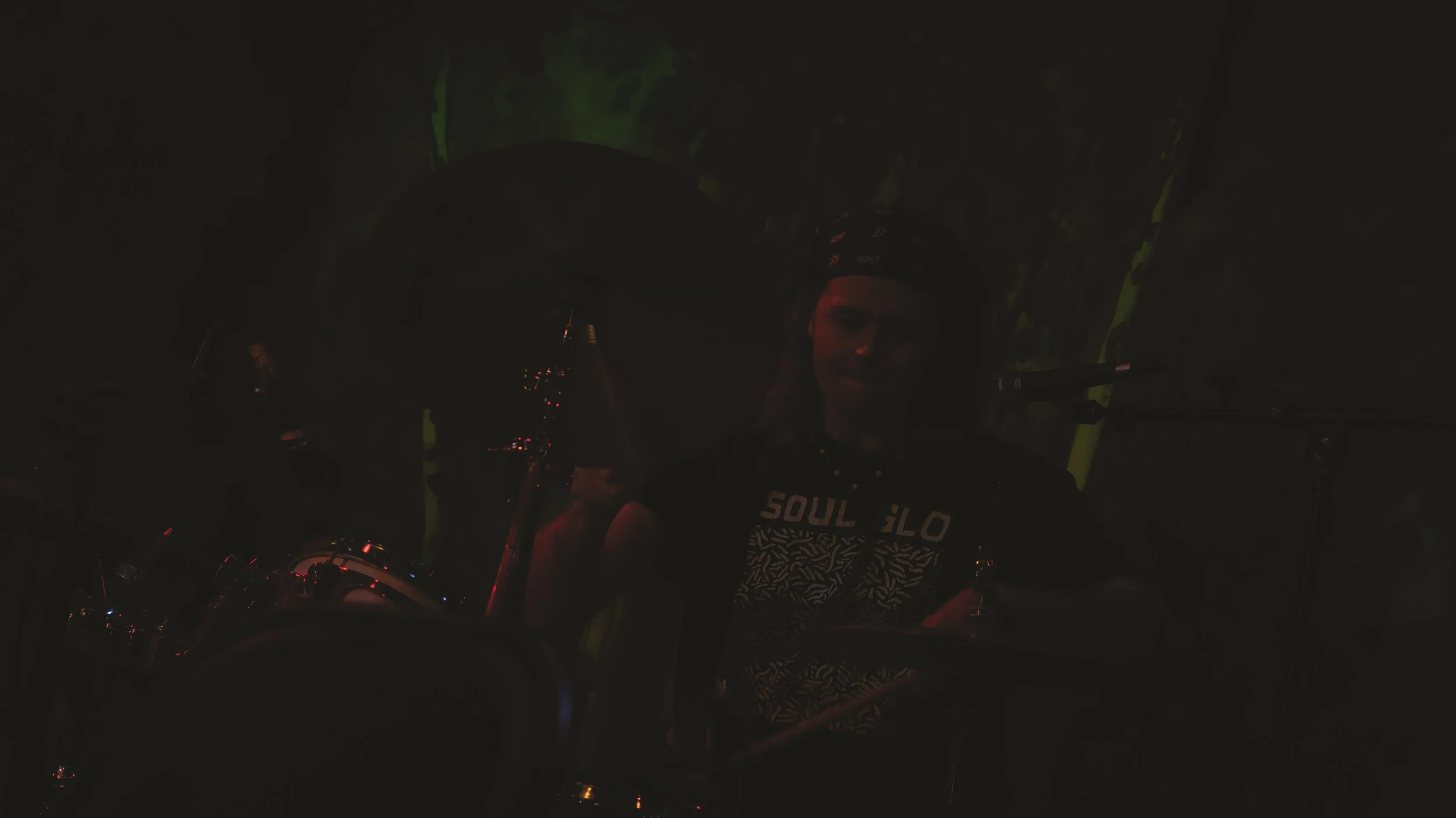 A person wearing a bandana and a black t-shirt with a graphic that says 'SOUL GLO' stands in a dark room lit by small lights, with a motorcycle in front and a person in the background working on something.