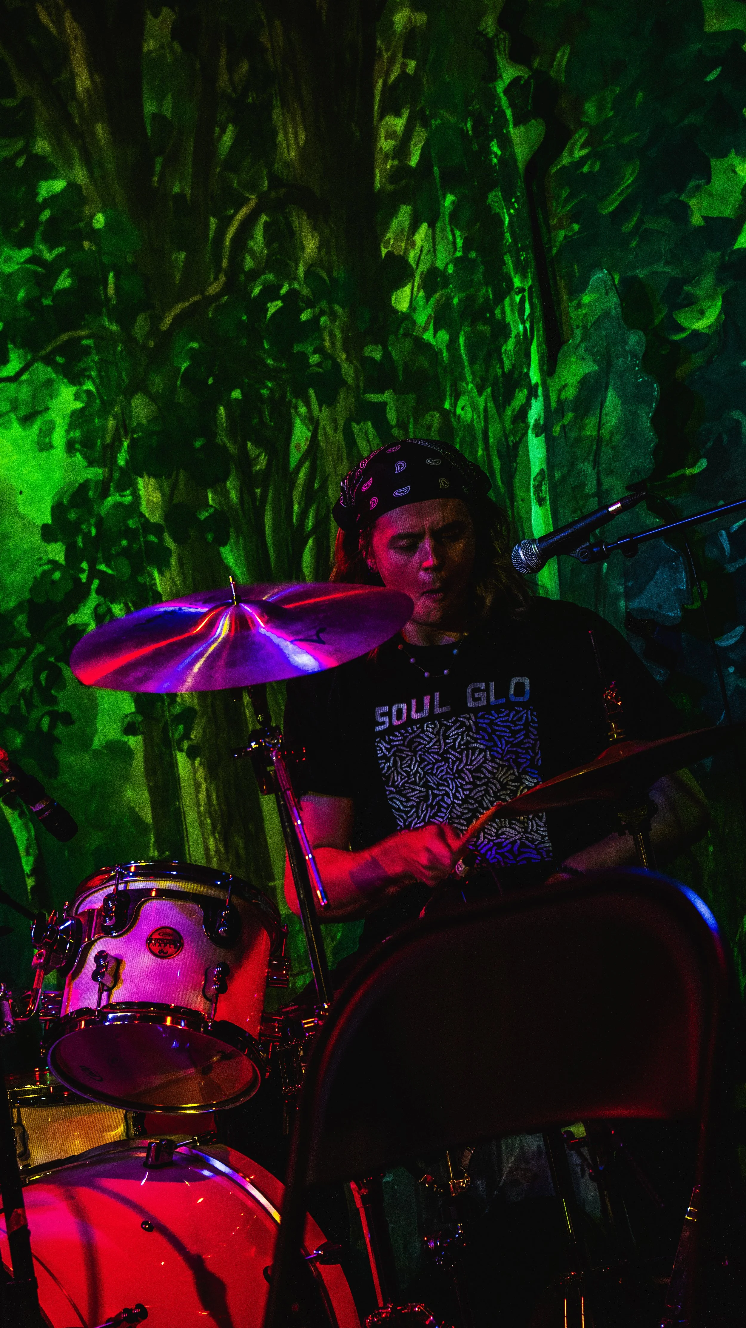 A woman playing drums on stage, wearing a bandana and a T-shirt that says 'Soul Glow,' with a green and earthy-toned mural in the background, illuminated by colorful stage lighting.