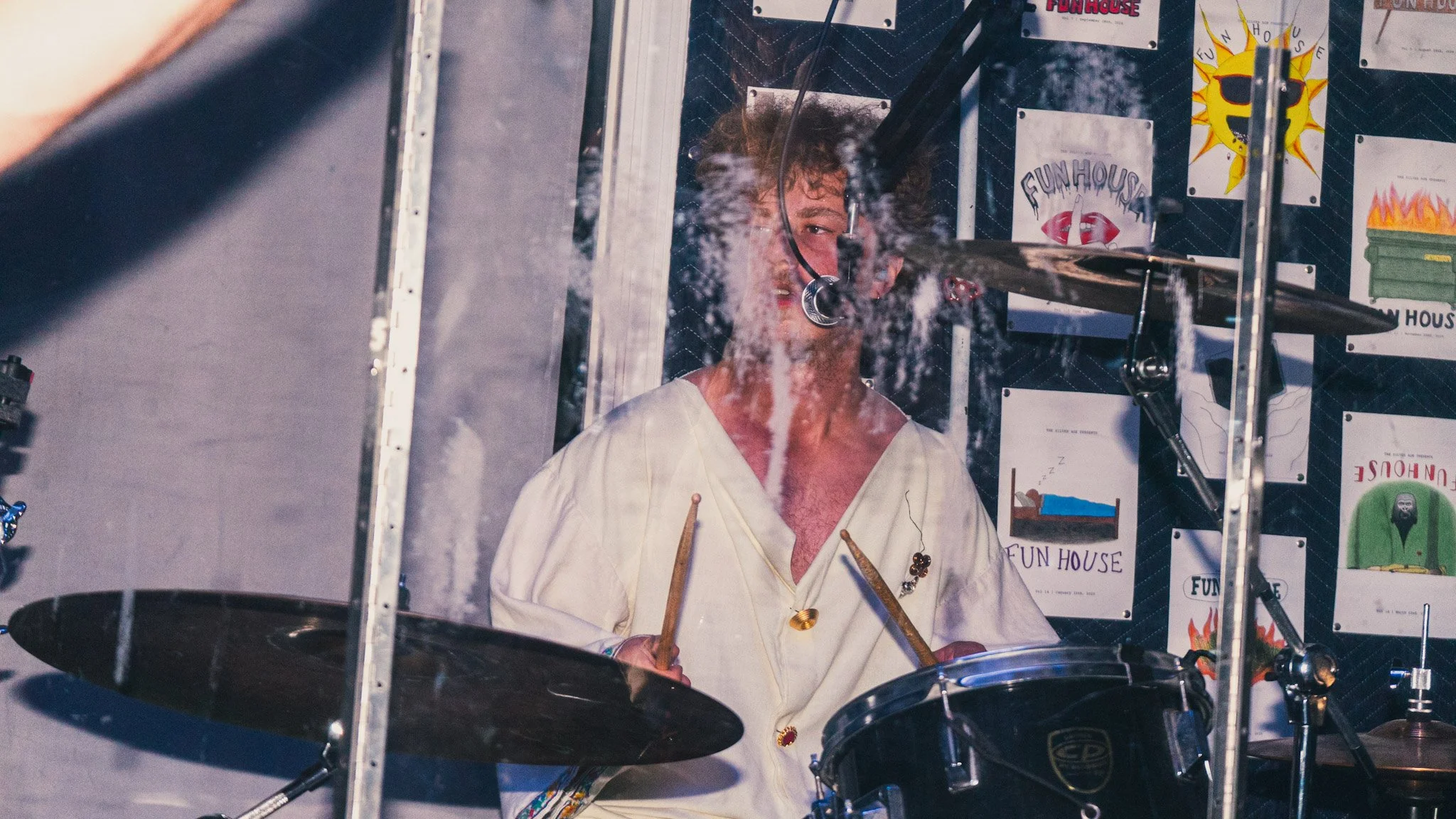 A person dressed as a drummer playing a drum set behind a glass or transparent barrier. The person has curly hair, is wearing a light-colored shirt, and is surrounded by colorful signs and posters with vibrant designs and text.