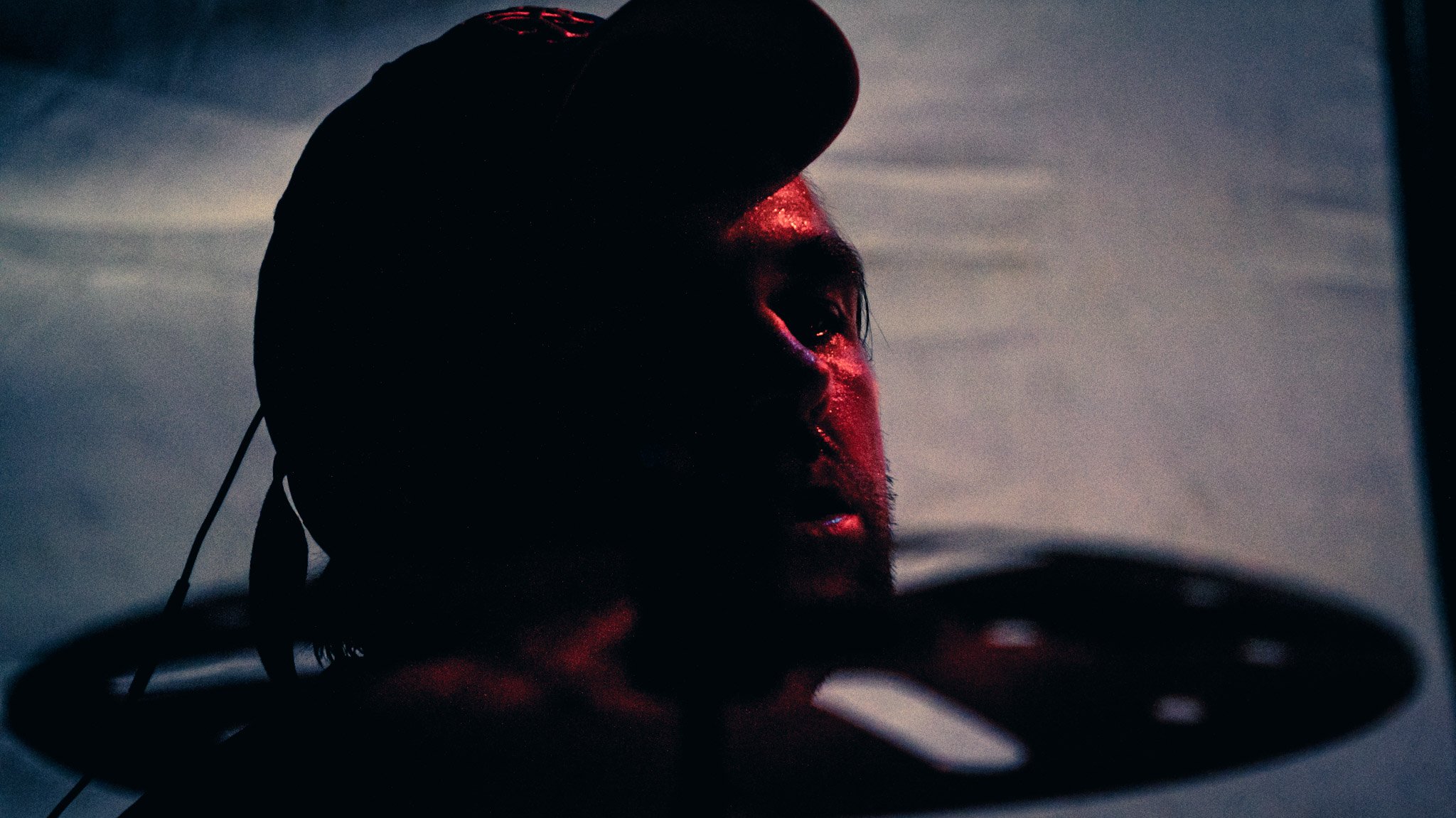A person wearing a cap with a curved brim and earphones, with their face partially illuminated by a red light, against a dark background with a cloudy sky.