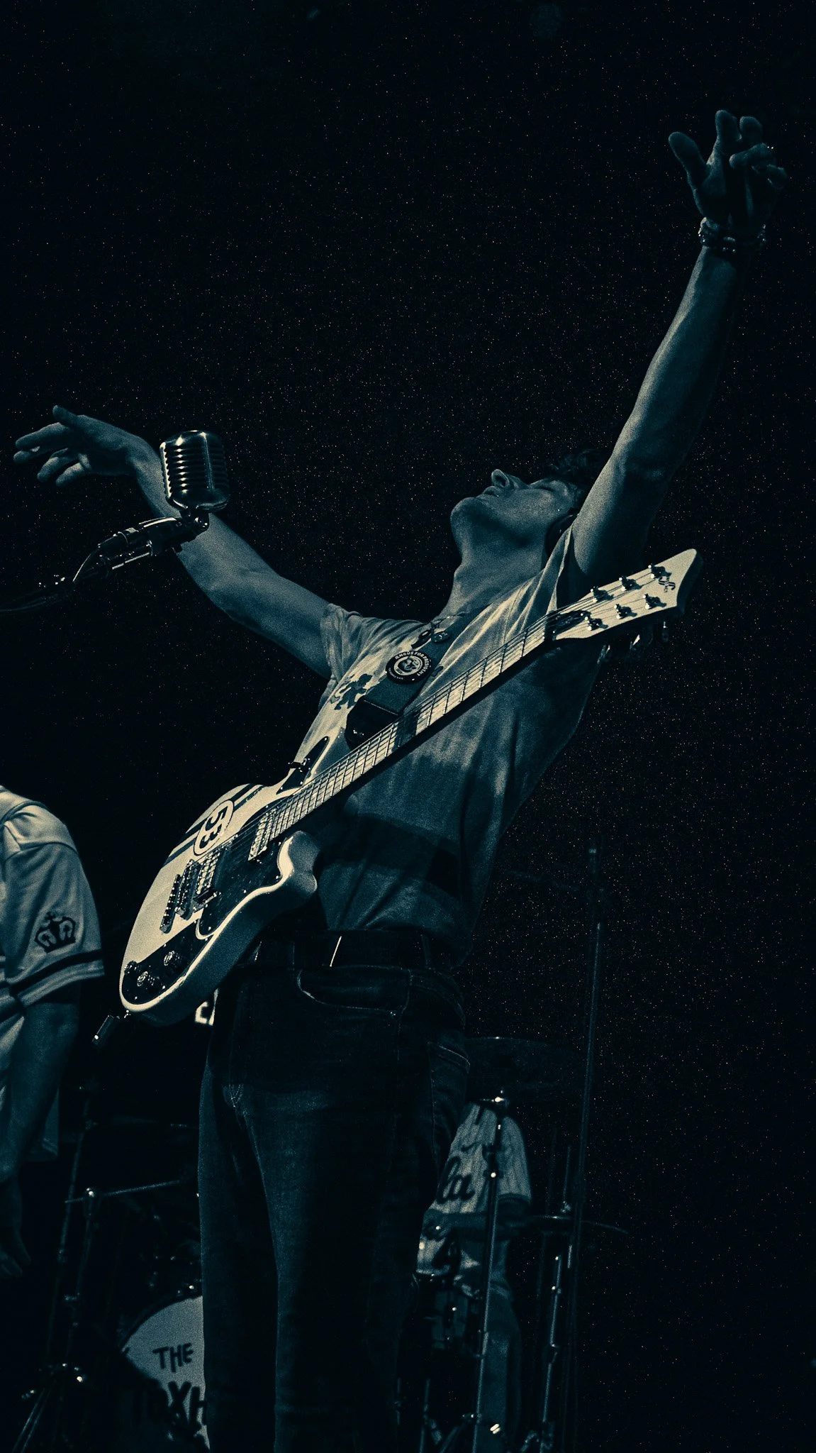 A musician dressed in a T-shirt with a guitar strap around his neck, performing with arms raised and a vintage microphone in front of him, against a starry night sky.