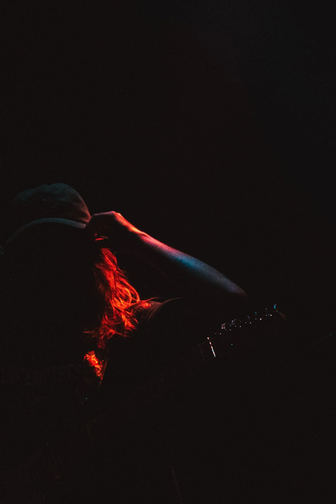 A person with long red hair, wearing a white hooded jacket and red pants, is sitting on the ground at night under a starry sky, illuminated by a red light.