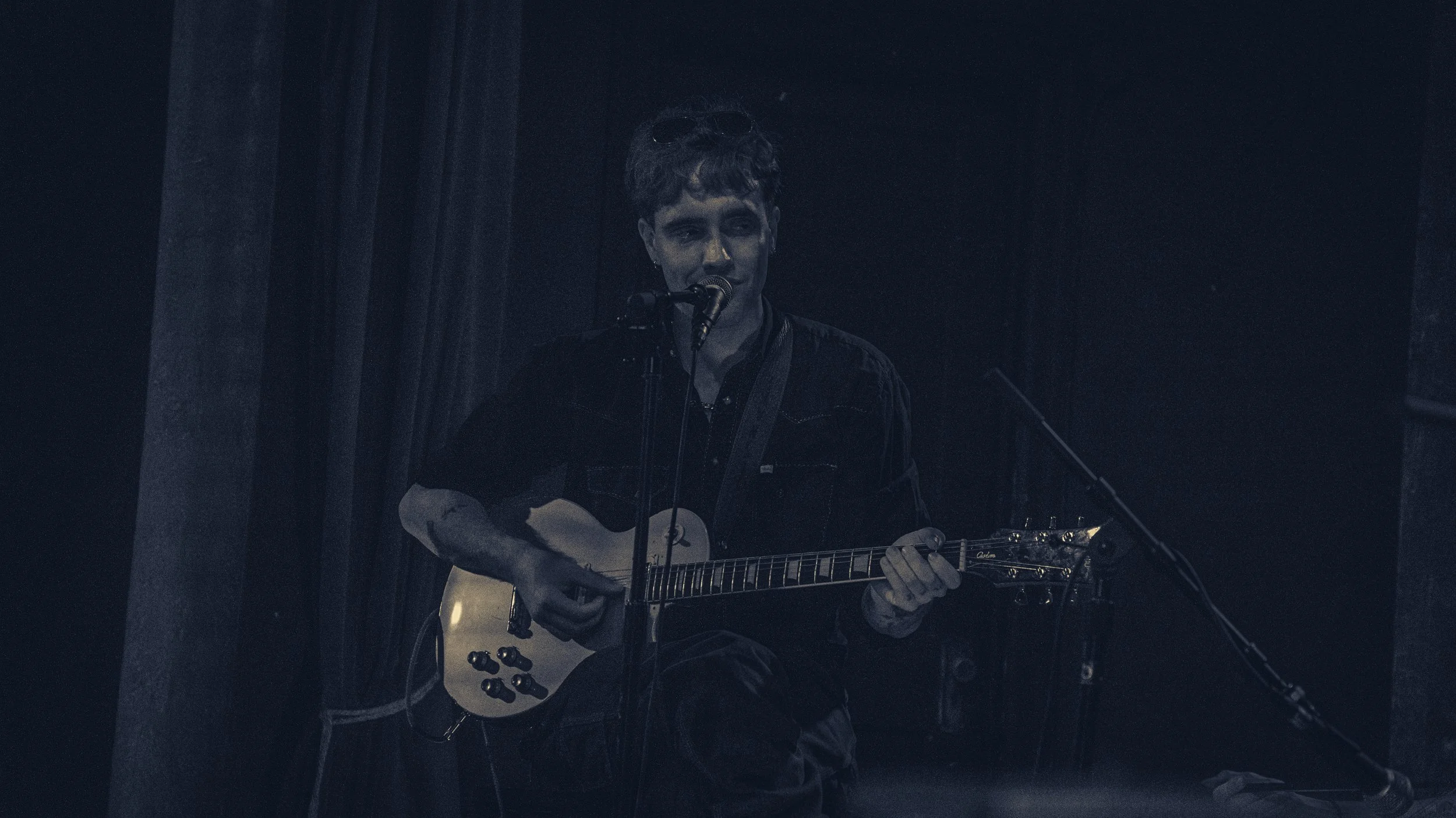 A person sitting on a stage playing an acoustic guitar and singing into a microphone, with dark curtains in the background.