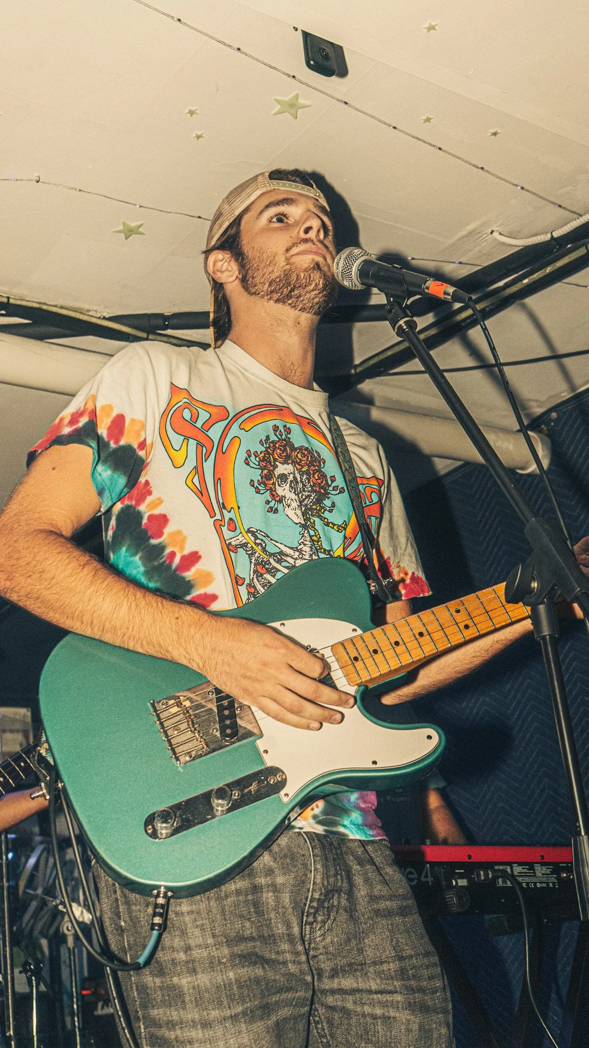 A man with a beard wearing a tie-dye T-shirt and a cap playing an electric guitar on stage, with a microphone in front of him.