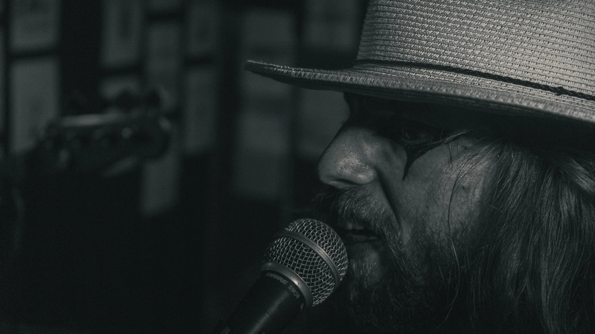 A close-up black-and-white photo of a person wearing a hat, singing or speaking into a microphone, with long hair partially visible.