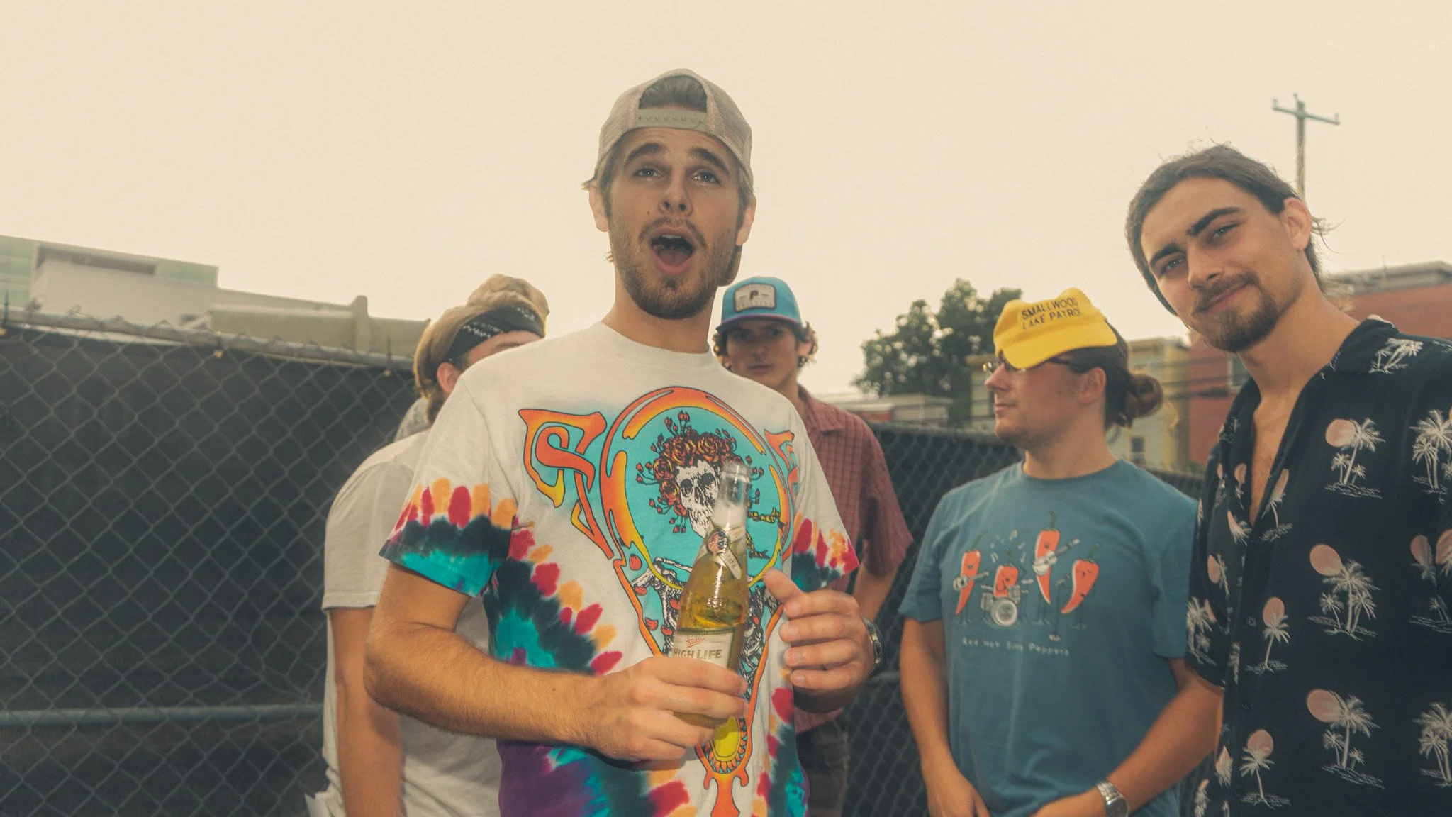 Group of young adults at an outdoor gathering, with one man in the foreground holding a beer, wearing a colorful tie-dye shirt, baseball cap backwards, and singing or speaking with his mouth open.