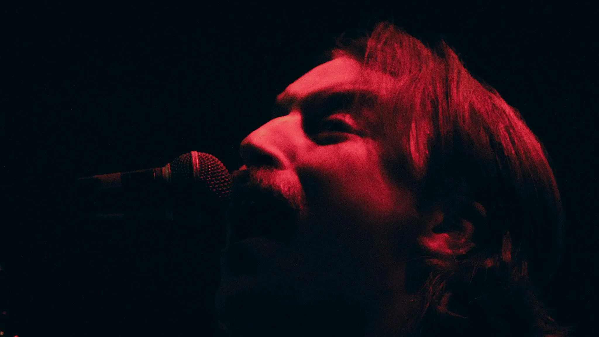 Close-up of a singer with long hair, singing into a microphone in a dark setting illuminated by red lighting.