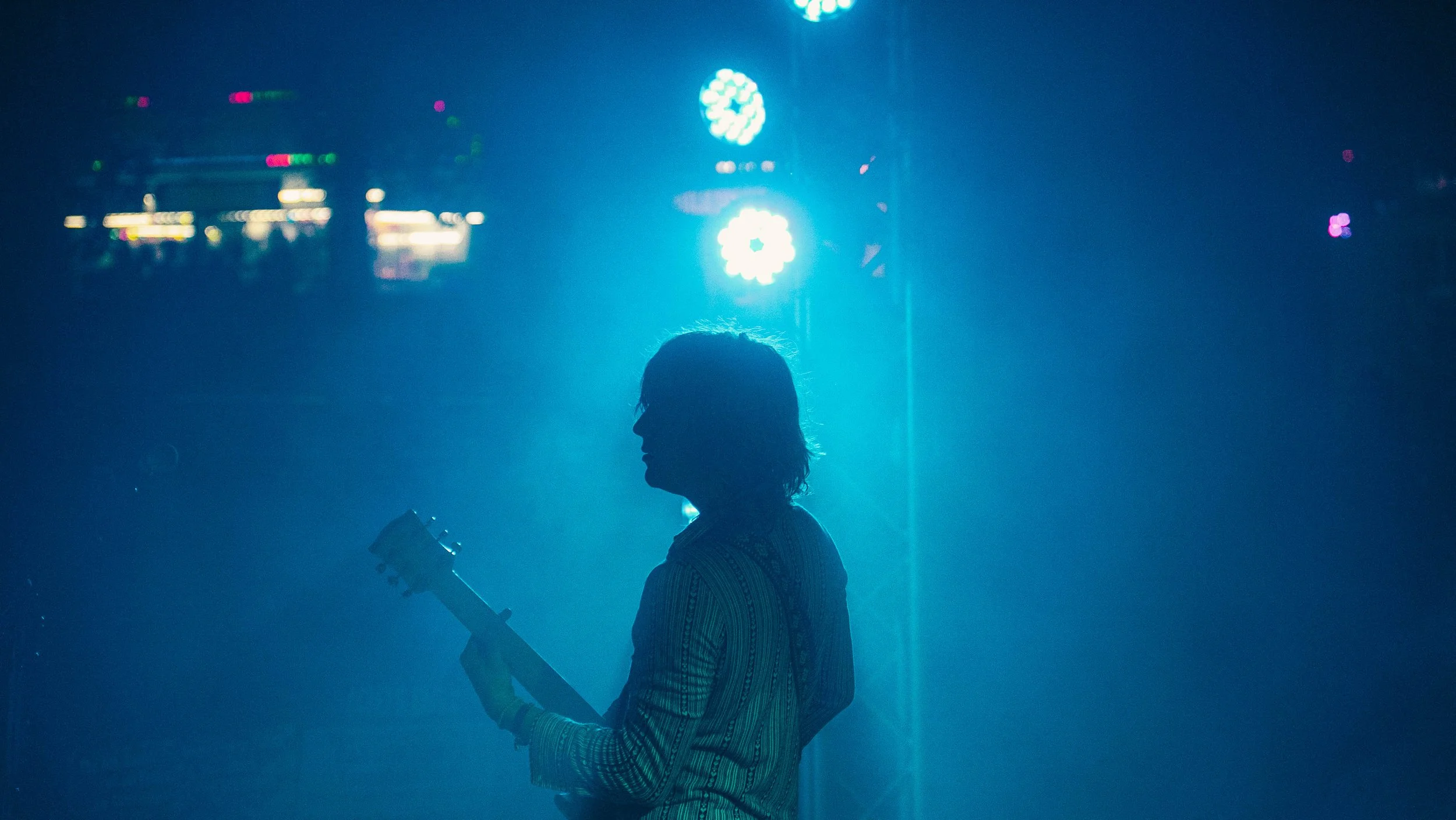 A person silhouettes while holding a guitar in front of bright stage lights at a concert or music event.