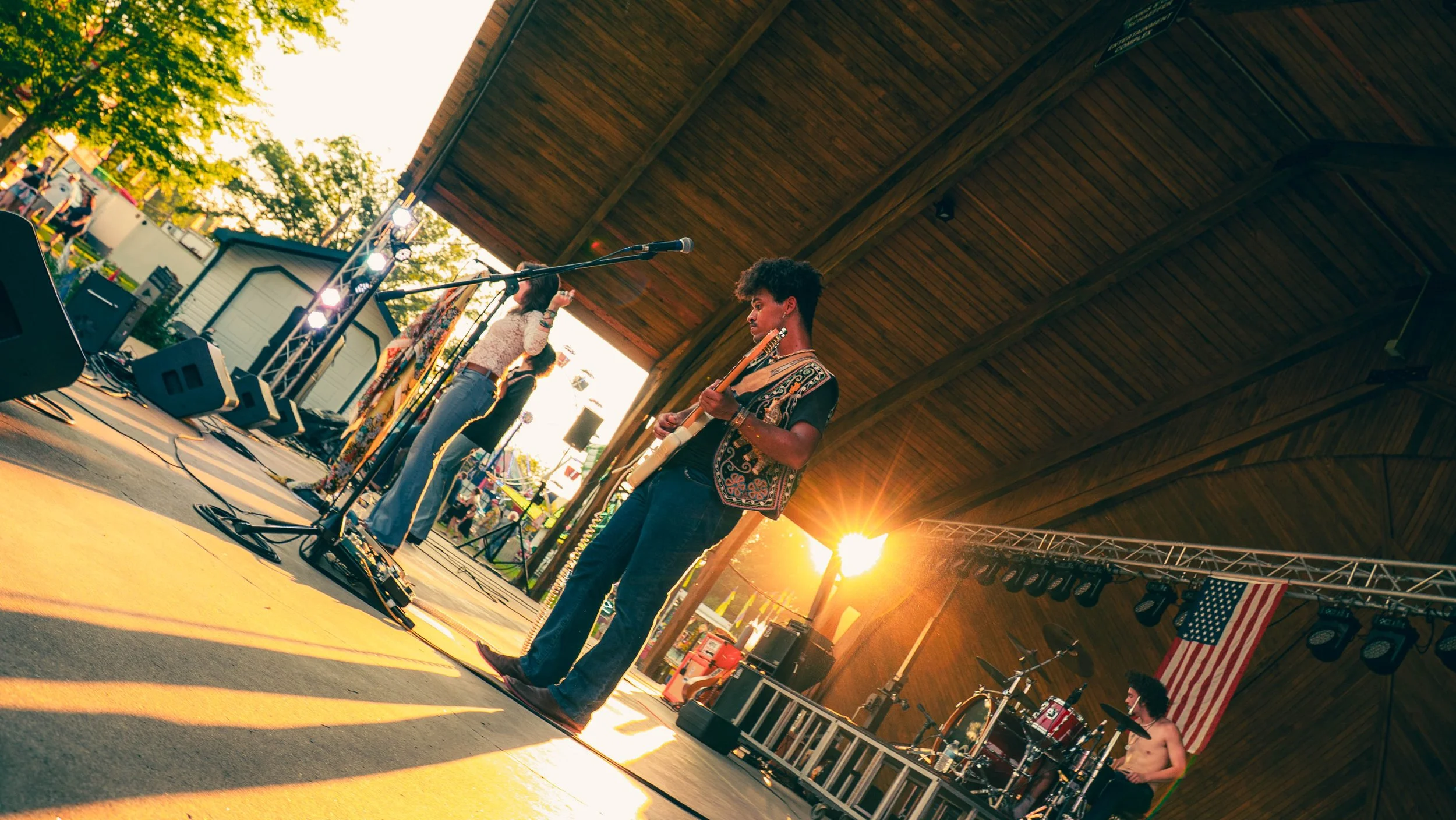 Musicians performing on outdoor stage at sunset, with the American flag hanging in the background.