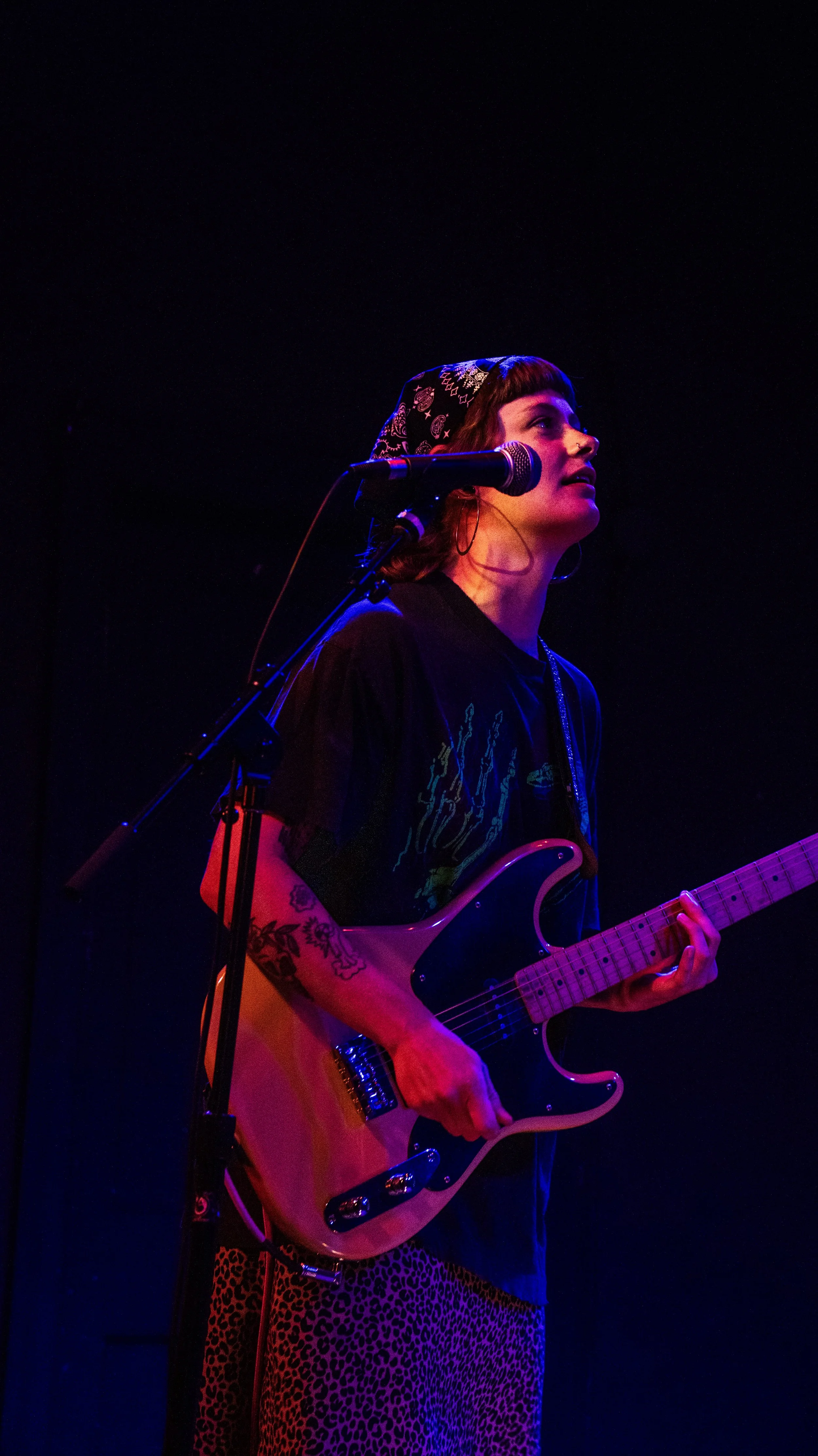A woman with tattoos on her arm, wearing a bandana and earrings, playing an electric guitar on stage with colorful stage lighting.