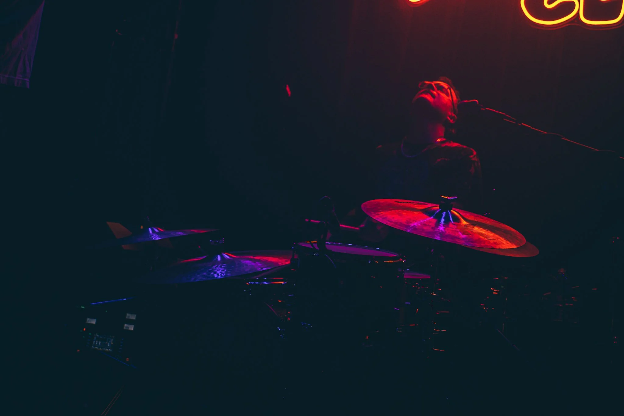A person performing on drums on a dark stage illuminated by red and purple neon lights.