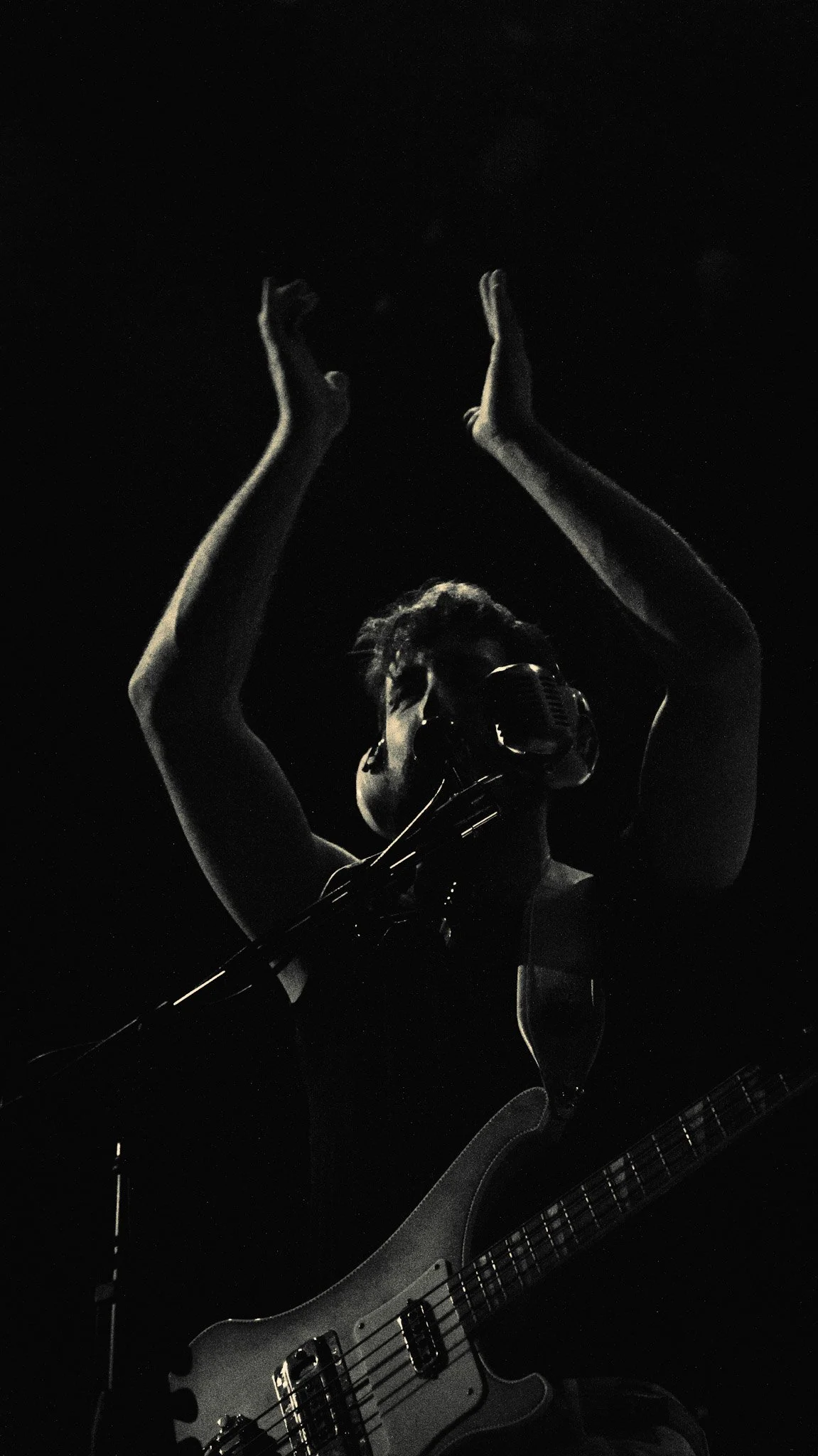 A person wearing glasses singing into a vintage microphone while playing an electric guitar in a dark setting.