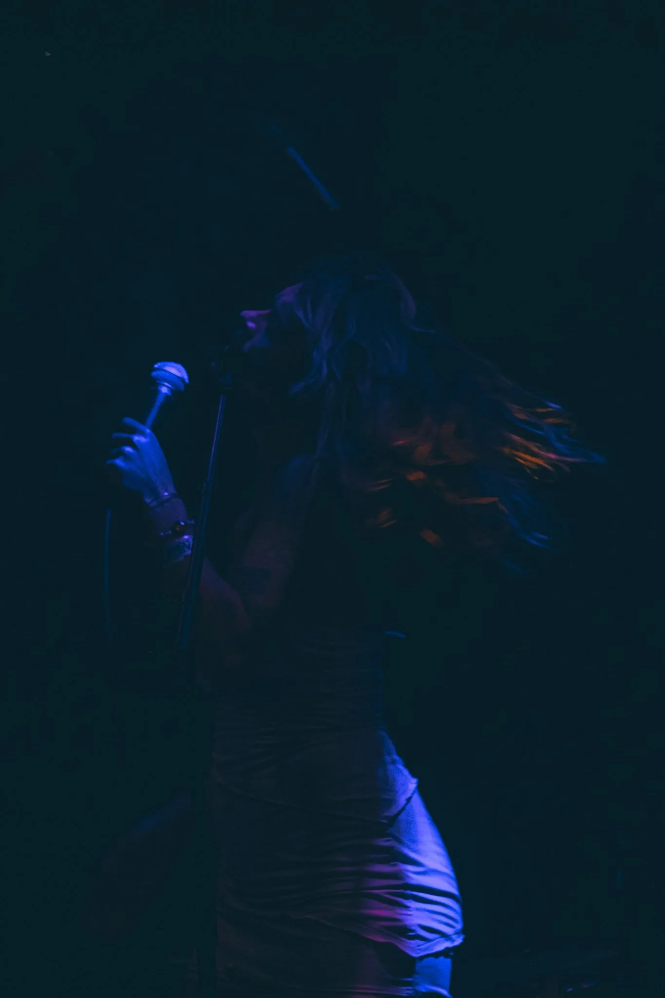 A female singer performing on stage under dim, colorful lighting, holding a microphone near her mouth, with her long hair flowing and wearing a white outfit.